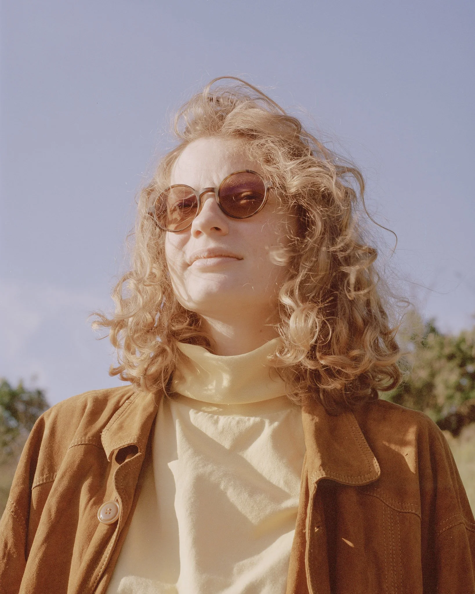 Nahaufnahme eines jungen Mannes mit lockigem, blondem Haar, trägt runde Sonnenbrillen und einen beige Pullover unter einem braunen Jacke, steht im Freien bei sonnigem Wetter.