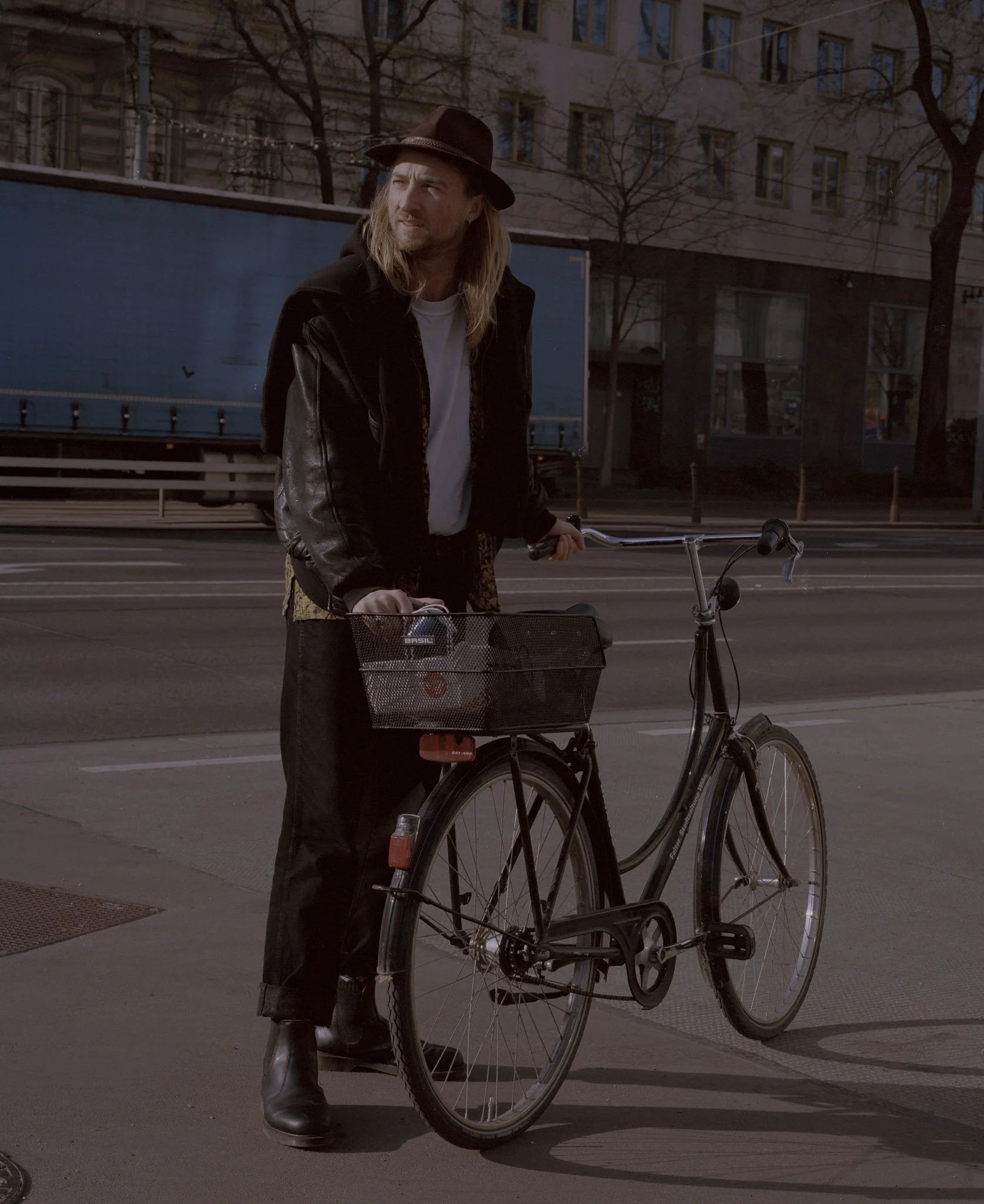 Mann mit langem Haar und Hut, steht neben Fahrrad auf Stadtstraße bei Dämmerung, im Hintergrund Gebäude und Bäume ohne Blätter.