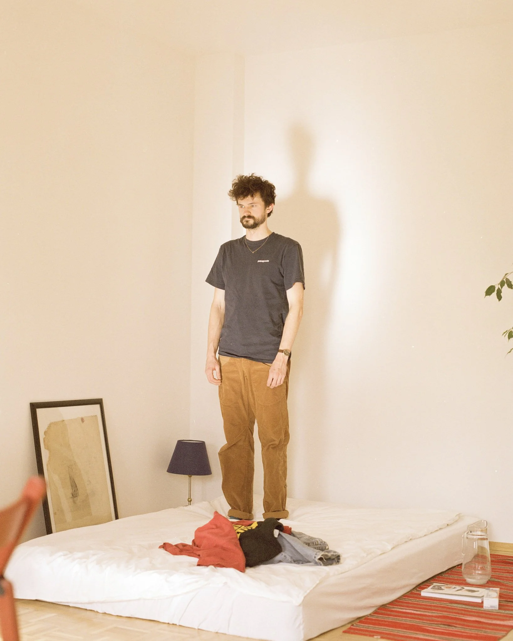 Ein Mann mit dunklem Haar, Bart und Mustache steht auf einem großen, ungemachten Bett in einem einfachen Raum. Er trägt ein schwarzes T-Shirt mit weißem Text und braune Hosen. Im Hintergrund ist die Wand hell, einige Bilder liegen auf dem Boden neben