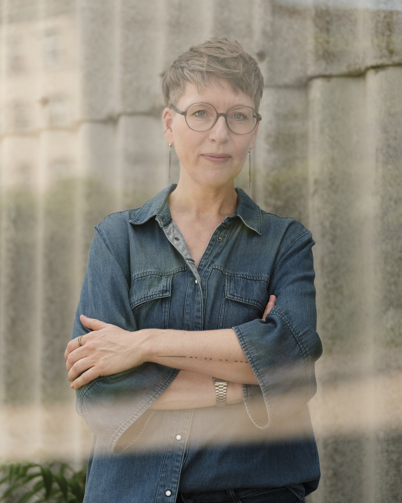 Eine Frau mit kurzen braunen Haaren, Brille, longen Armbändern und Ohrringen, trägt eine Jeansjacke und eine beige Hose. Sie steht vor einer Steinmauer mit ihren Armen verschränkt.