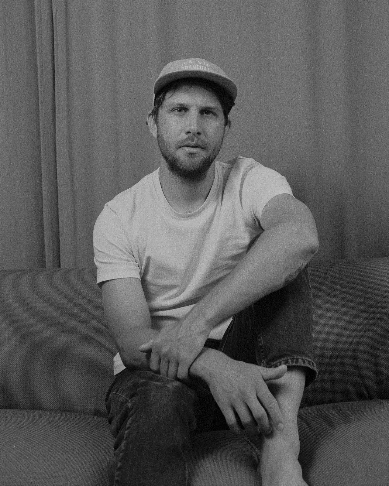 Schwarz-weiß-Foto eines Mannes mit Bart und Kurzhaar, trägt ein Baseballcap und ein T-Shirt, sitzt auf einem Sofa in einem Raum mit Holzvertäfelung im Hintergrund.