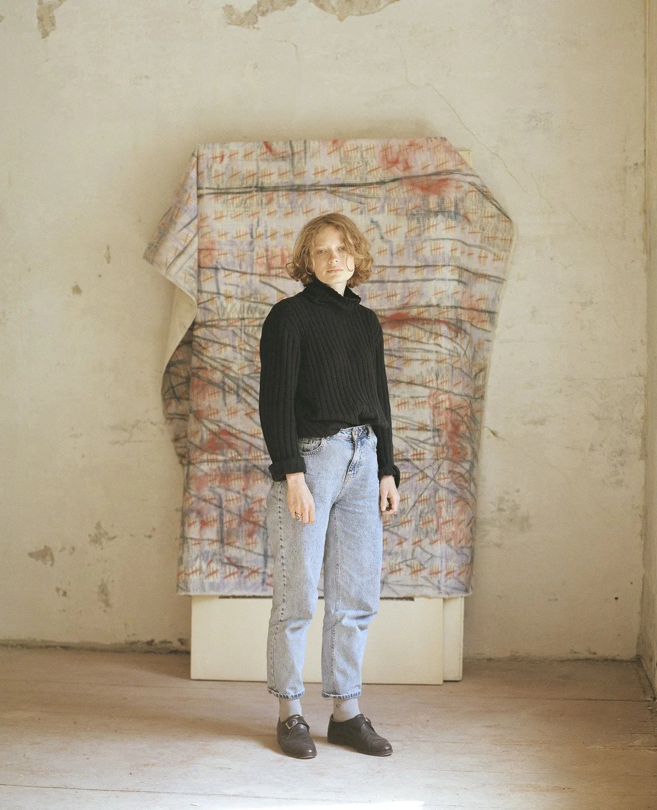 Eine junge Frau steht vor einer bemalten Wand in einem abgenutzten Raum, trägt einen schwarzen Pullover, helle Jeans und lässige Schuhe.