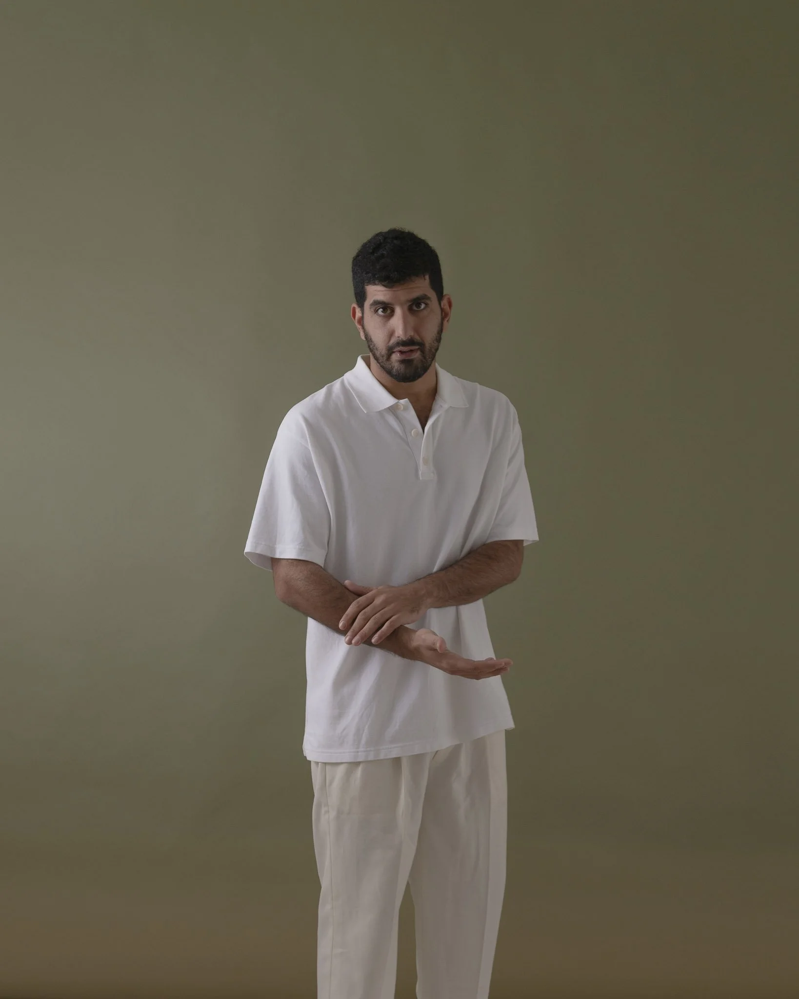 Ein Mann mit dunklem Haar und Bart in weißem Polo und weißen Hosen steht vor einem grünen Hintergrund, die Arme verschränkt, Blick in die Kamera.