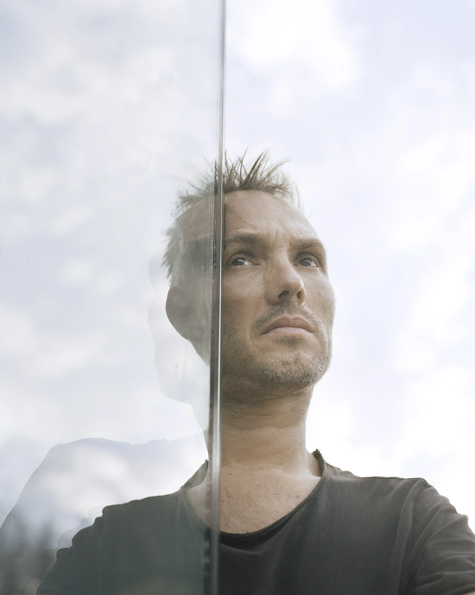 Ein Mann schaut durch eine Glaswand nach oben, seine Haare sind zerzaust, die Kleidung ist schwarz, der Himmel ist überwiegend bewölkt.