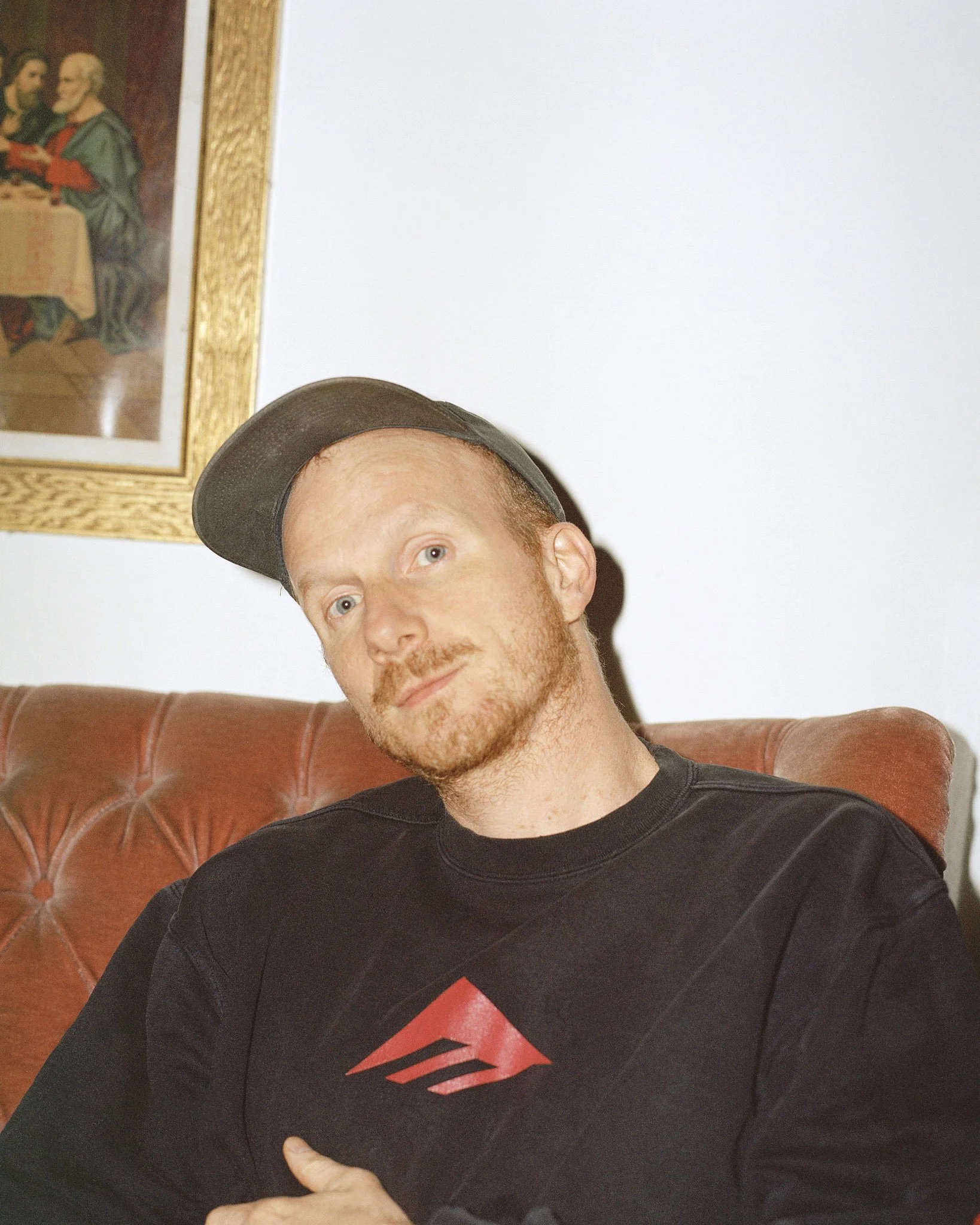 Junger Mann sitzt auf einem roten Ledersofa, trägt ein schwarzes T-Shirt mit einem roten Superman-Logo und eine schwarze Baseballmütze. Im Hintergrund ist ein Bild an der Wand zu sehen.