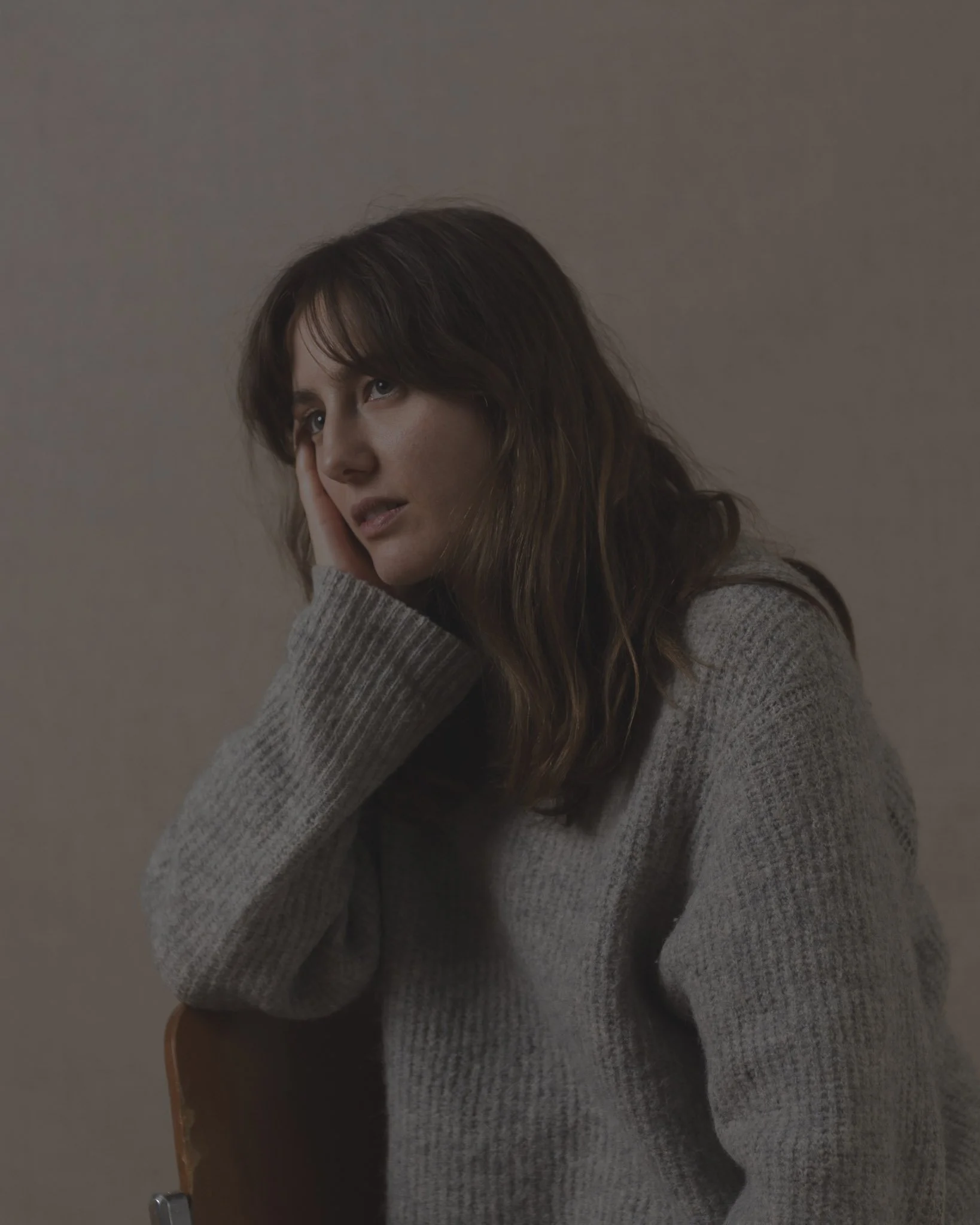 A woman with long brown hair wearing a gray sweater, resting her head on her hand, sitting against a plain wall.