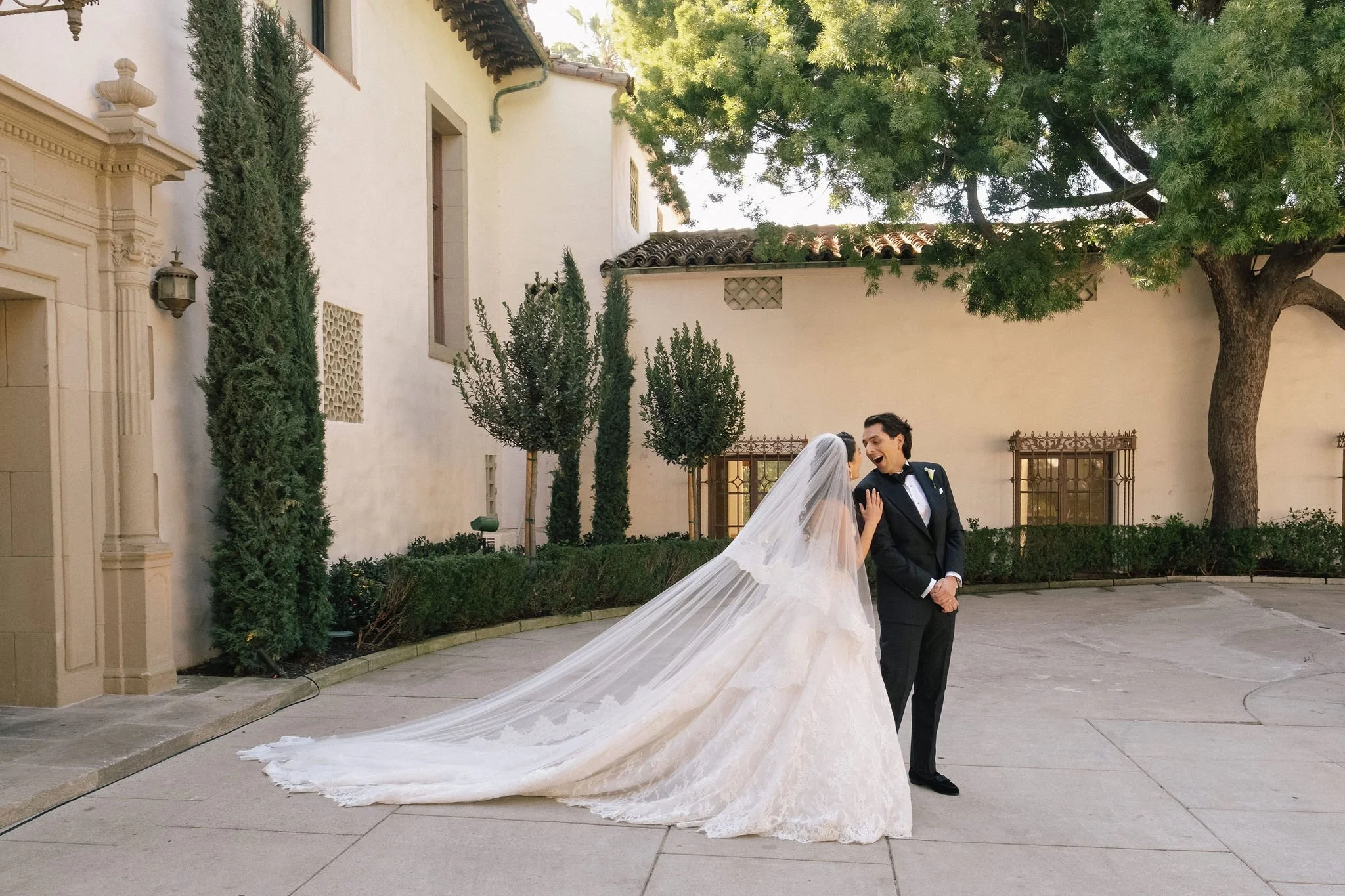 A bride in a wedding dress and veil is laughing and touching a groom in a tuxedo in an outdoor courtyard with trees and bushes.