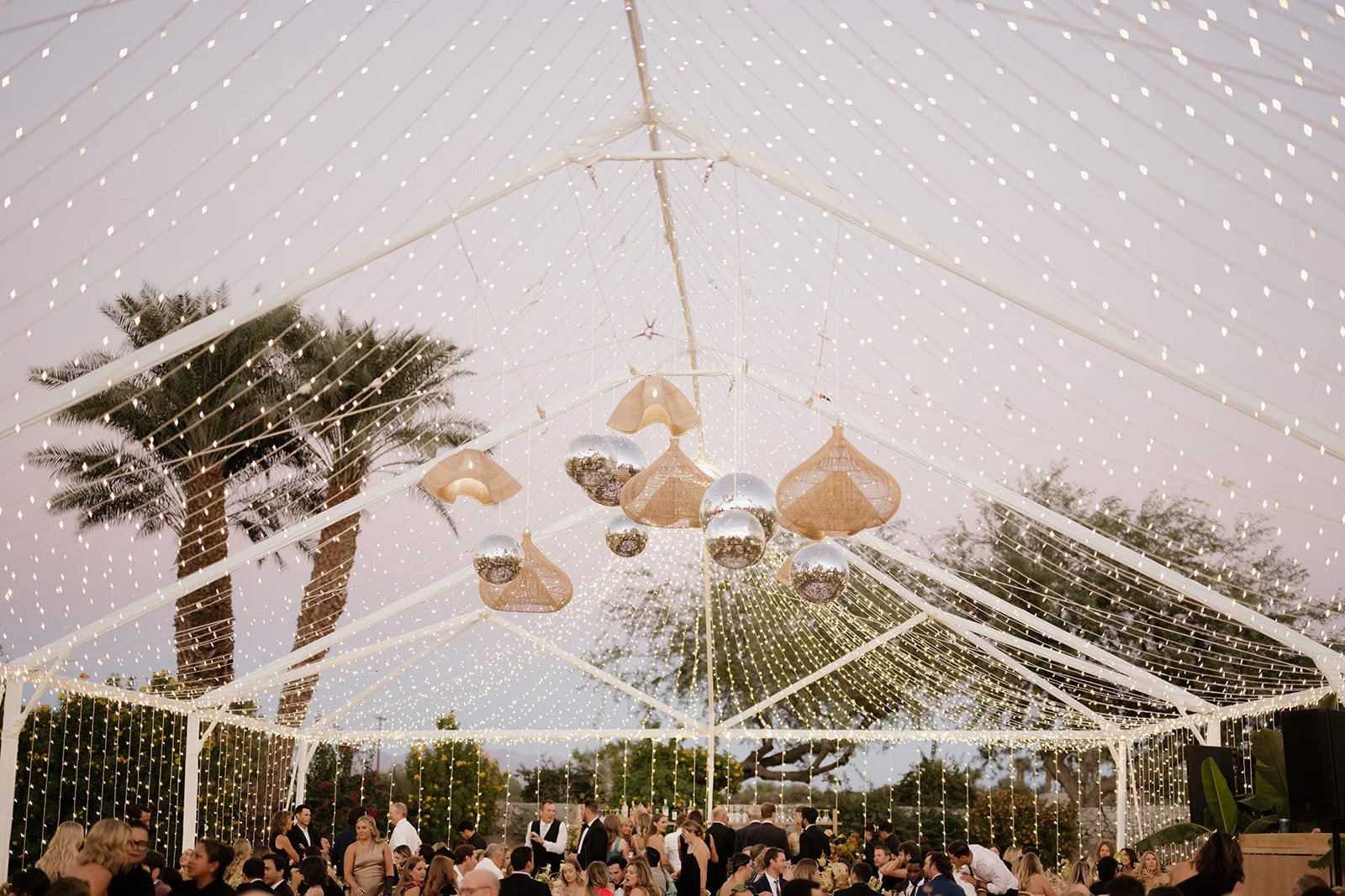An outdoor event under a white tent decorated with string lights and hanging metallic and wicker pendant lights, with palm trees and other trees in the background. There are many people attending the event.