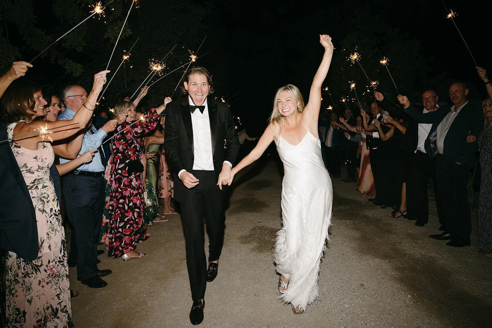 A newlywed couple holding hands and running through a path at night, surrounded by friends and family holding sparklers.