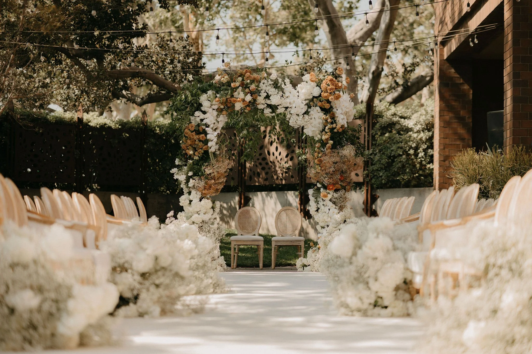 Outdoor wedding aisle decorated with white flowers, leading to a floral arch with white and peach blossoms. Two chairs sit beneath the arch, with string lights overhead and trees and buildings in the background.