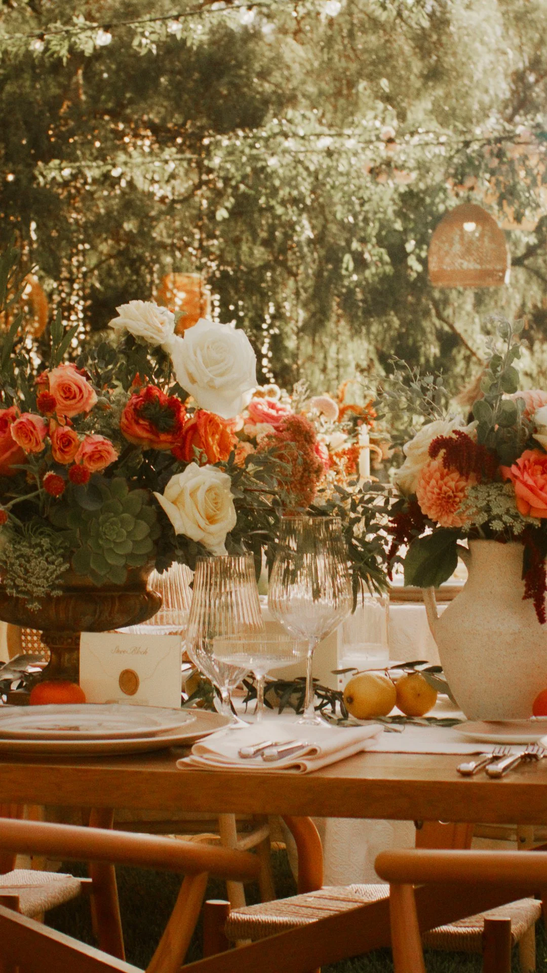 Decorated outdoor dining table with floral centerpieces, glassware, plates, cutlery, and lemons, set against a leafy background.