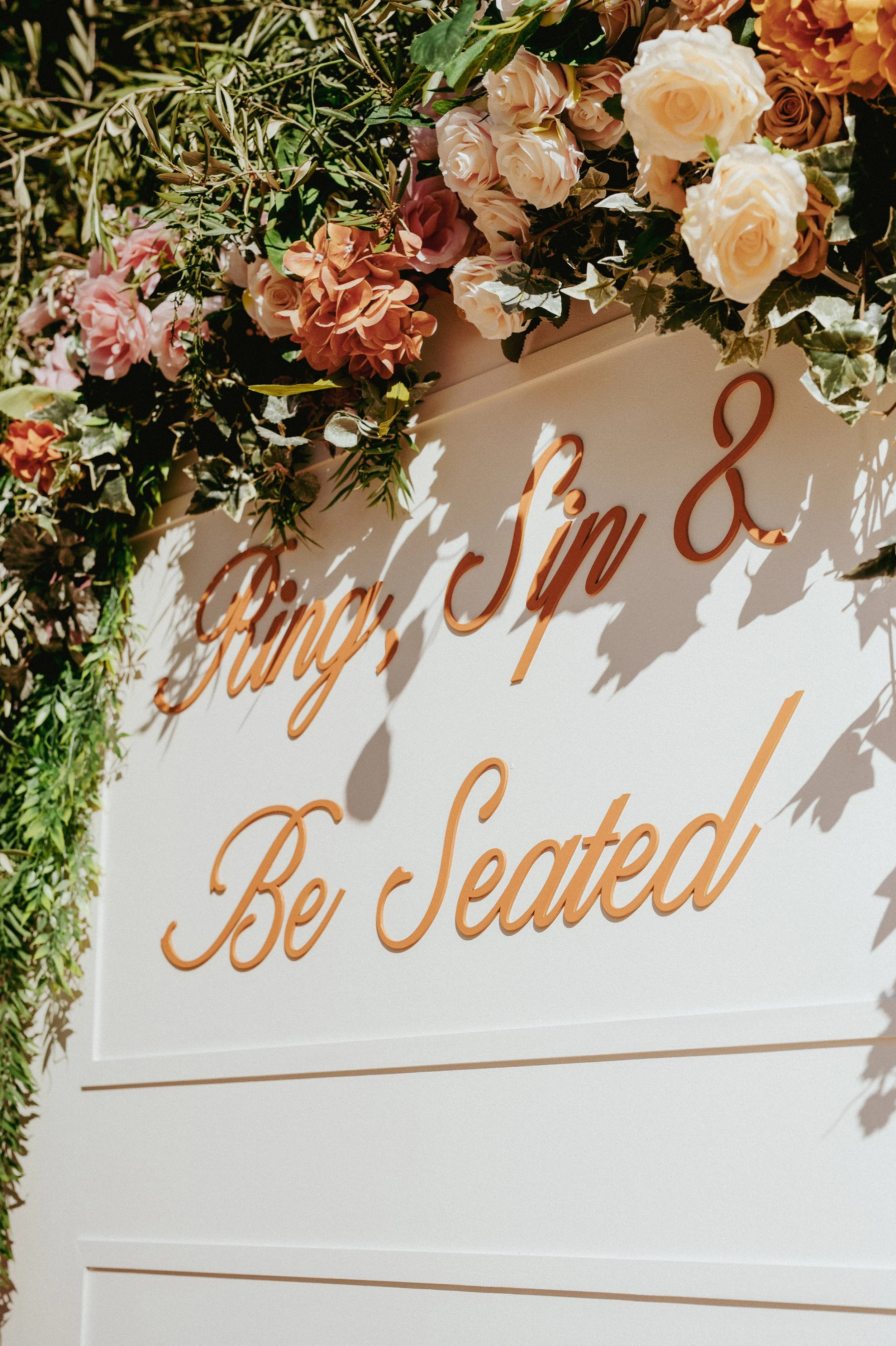 A drink wall lined with flowers that says "ring, sip and be seated"