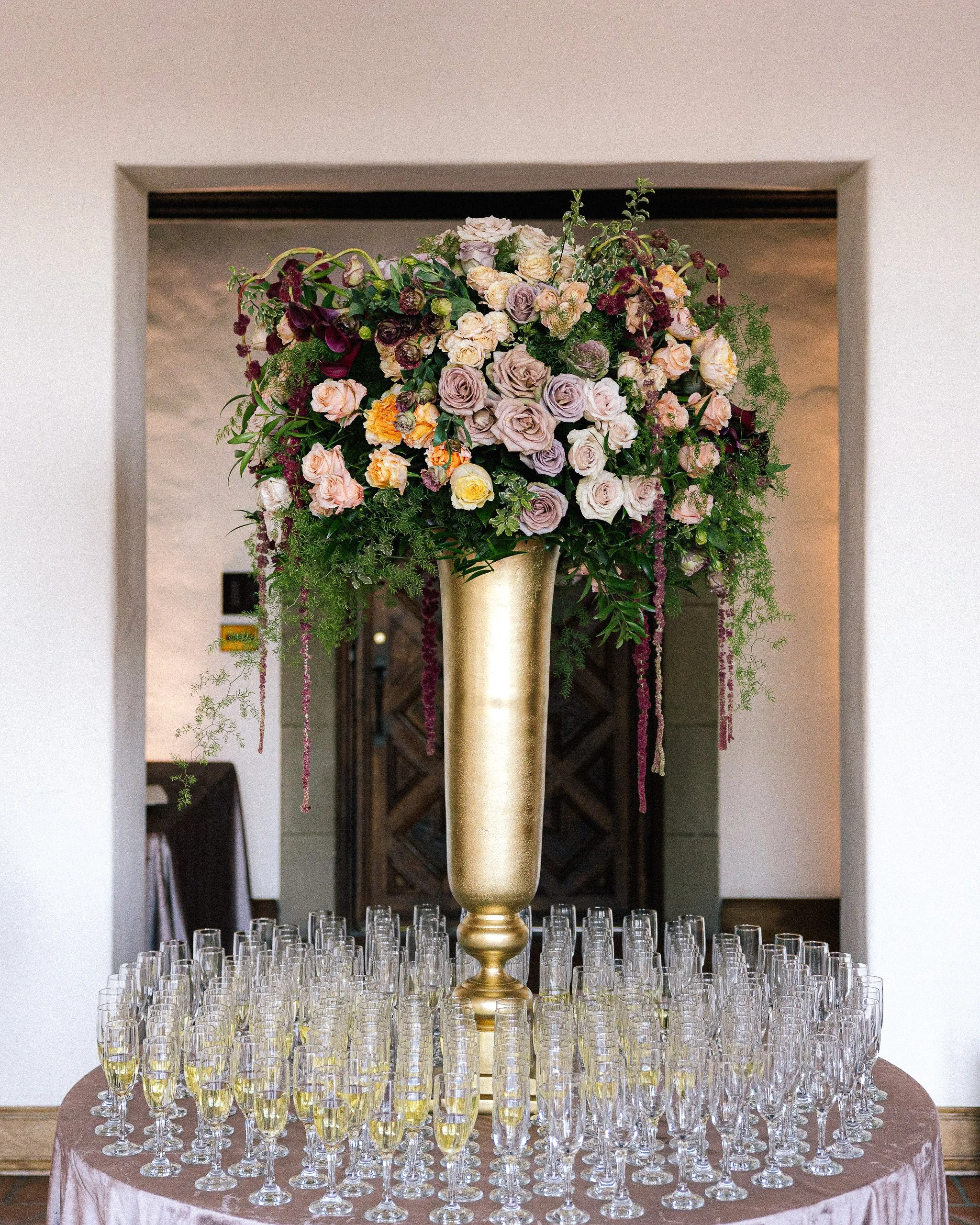 A large bouqet of flowers in a large gold vase surrounded by 75 or so champange glasses at a wedding