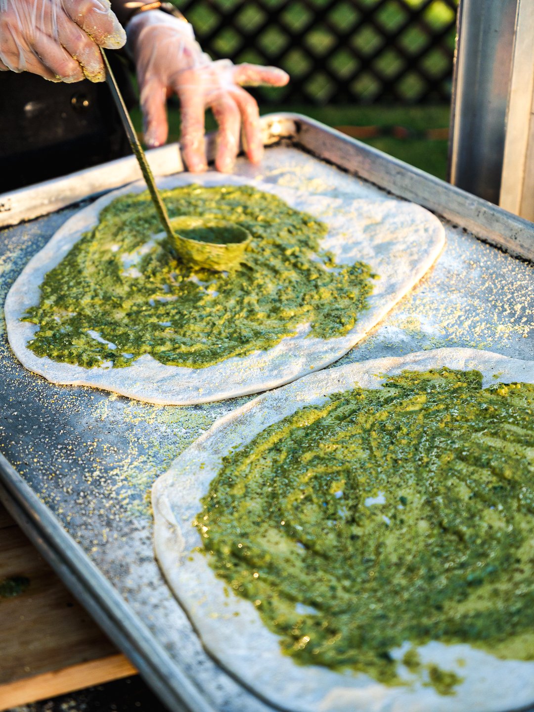 Hands spreading green pesto on pizza dough rounds on a baking sheet.