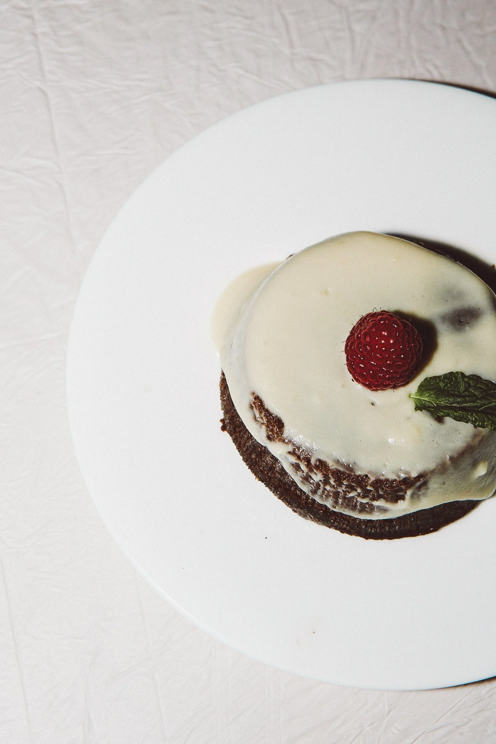 Chocolate cake with white frosting, topped with a raspberry and a mint leaf, on a white plate.