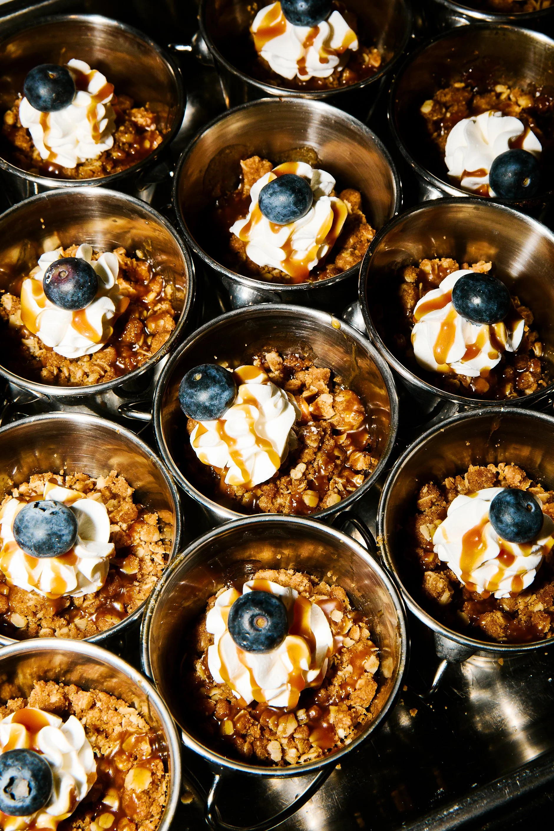 Multiple dessert cups with whipped cream, blueberries, caramel sauce, and crumbly topping.