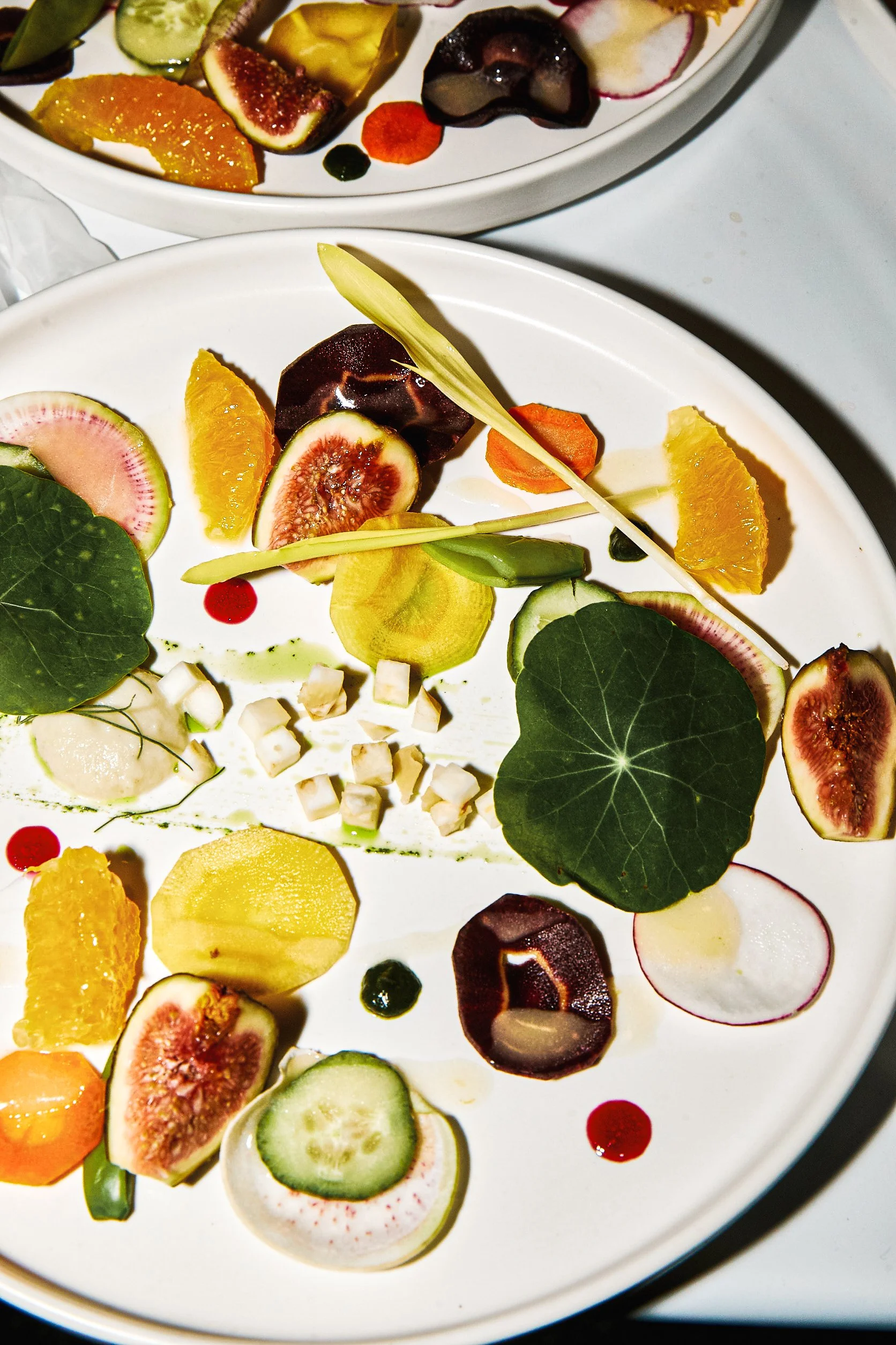 TGIS Catering Best Appetizer, plate of assorted fresh fruit and edible flowers, including figs, cucumber, radish, carrot, orange, and yellow fruit, garnished with small decorative cubes and sauce dots.