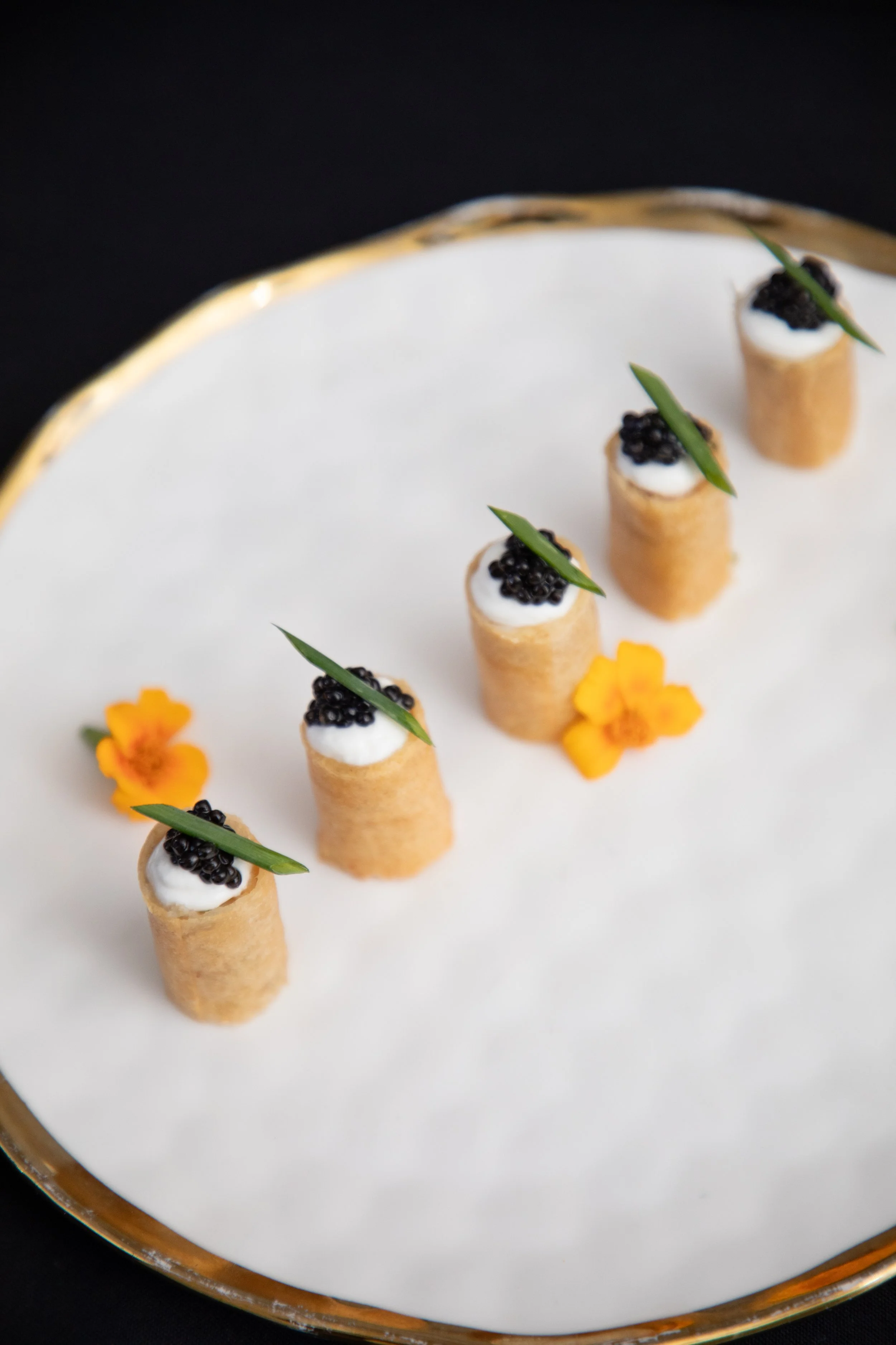 Five mini savory appetizers with pastry shells topped with black caviar, a small green garnish, and a dollop of cream, garnished with yellow edible flowers, arranged on a white and gold-rimmed plate.