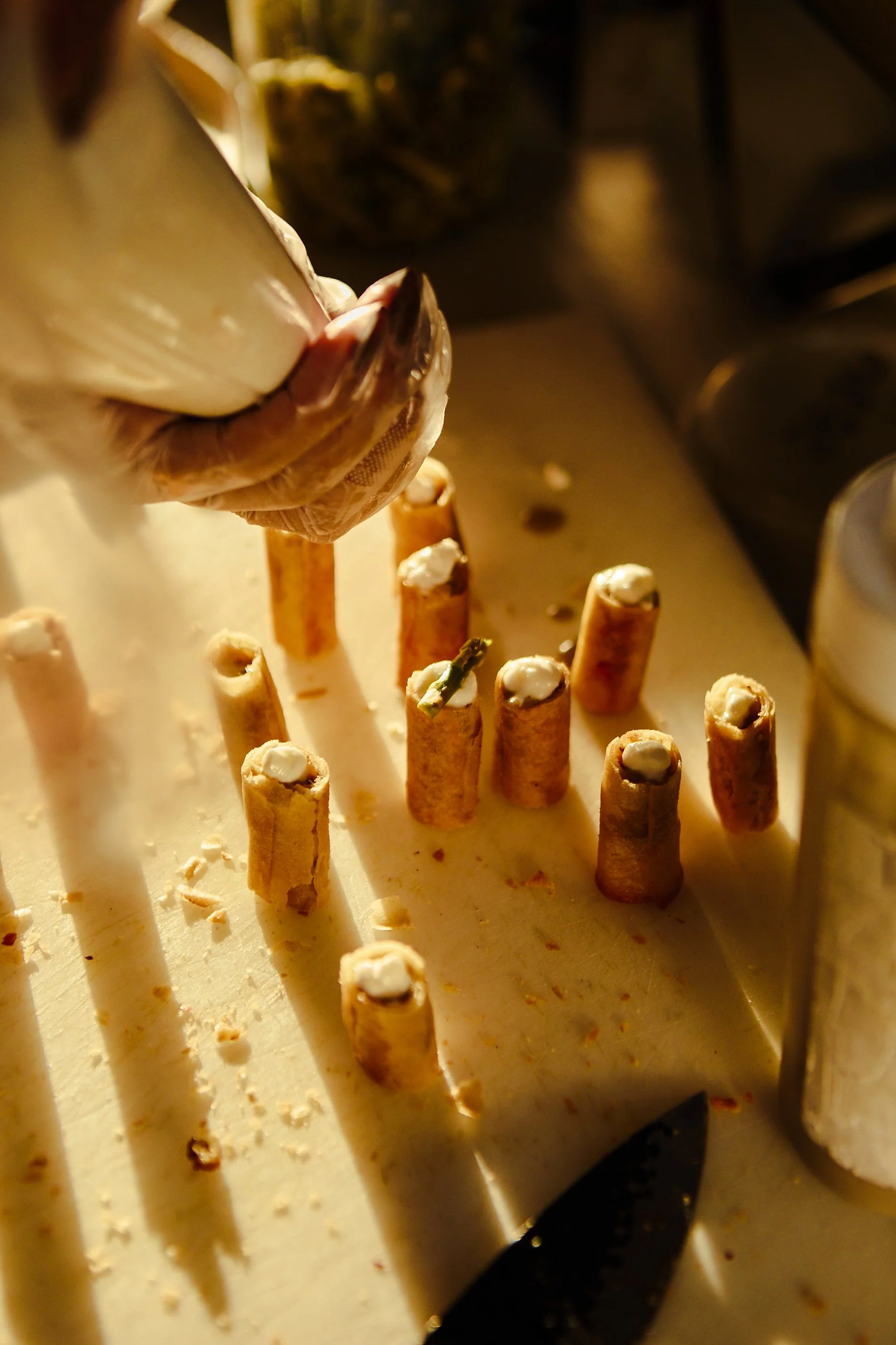 A person wearing gloves is filling baked pastry shells with a creamy filling. There are several empty shells on a tray, and a knife is visible at the bottom of the image.