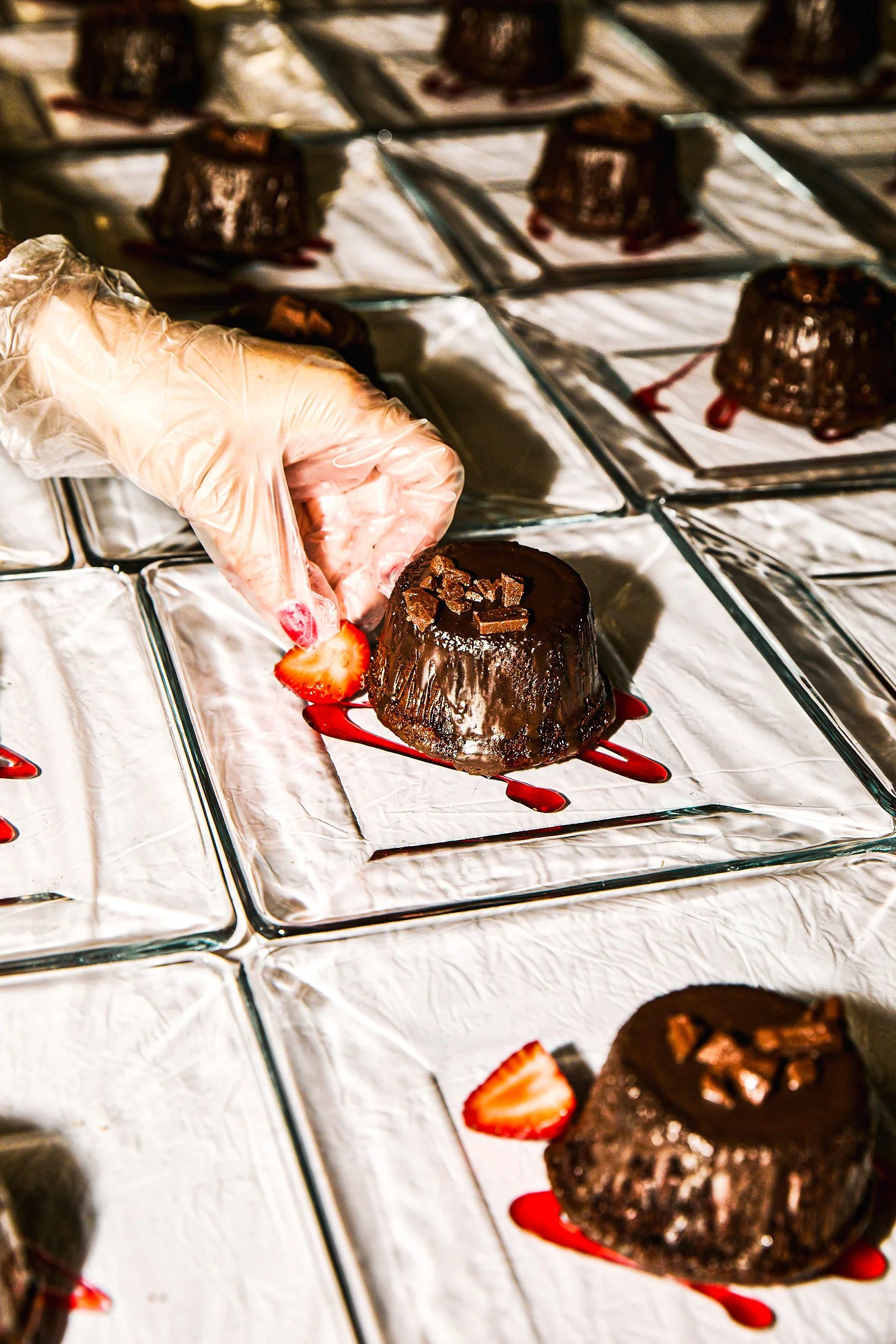 A person wearing a plastic glove adds a sliced strawberry to a chocolate mousse dessert on a silver tray, with other similar desserts in the background.
