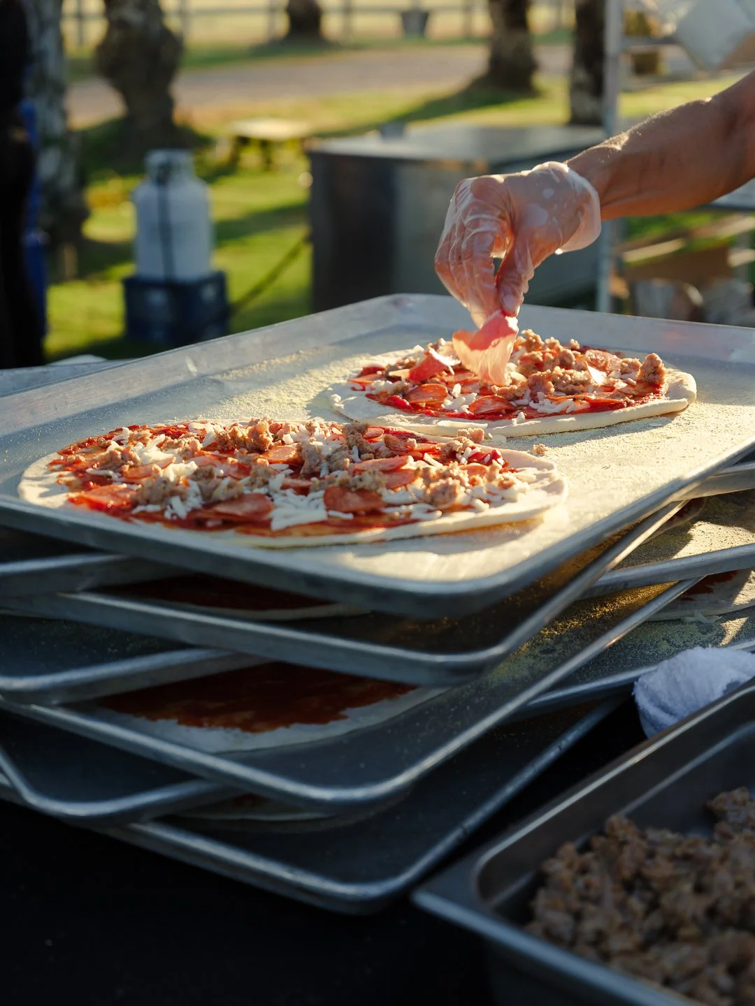 Person preparing pizza outdoors, arranging toppings on two uncooked pizzas on baking trays with a hand wearing a plastic glove.