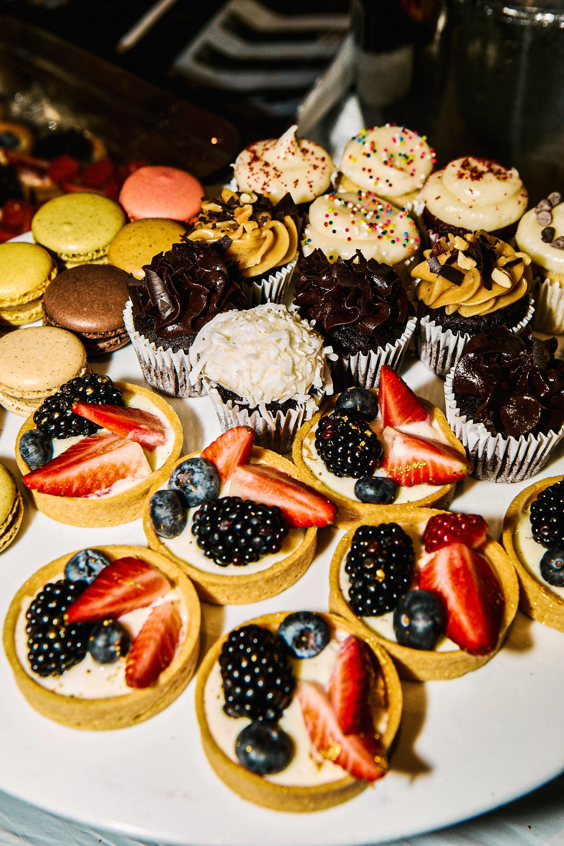 Assorted cupcakes, macarons, and fruit tarts on a white platter at a dessert table.
