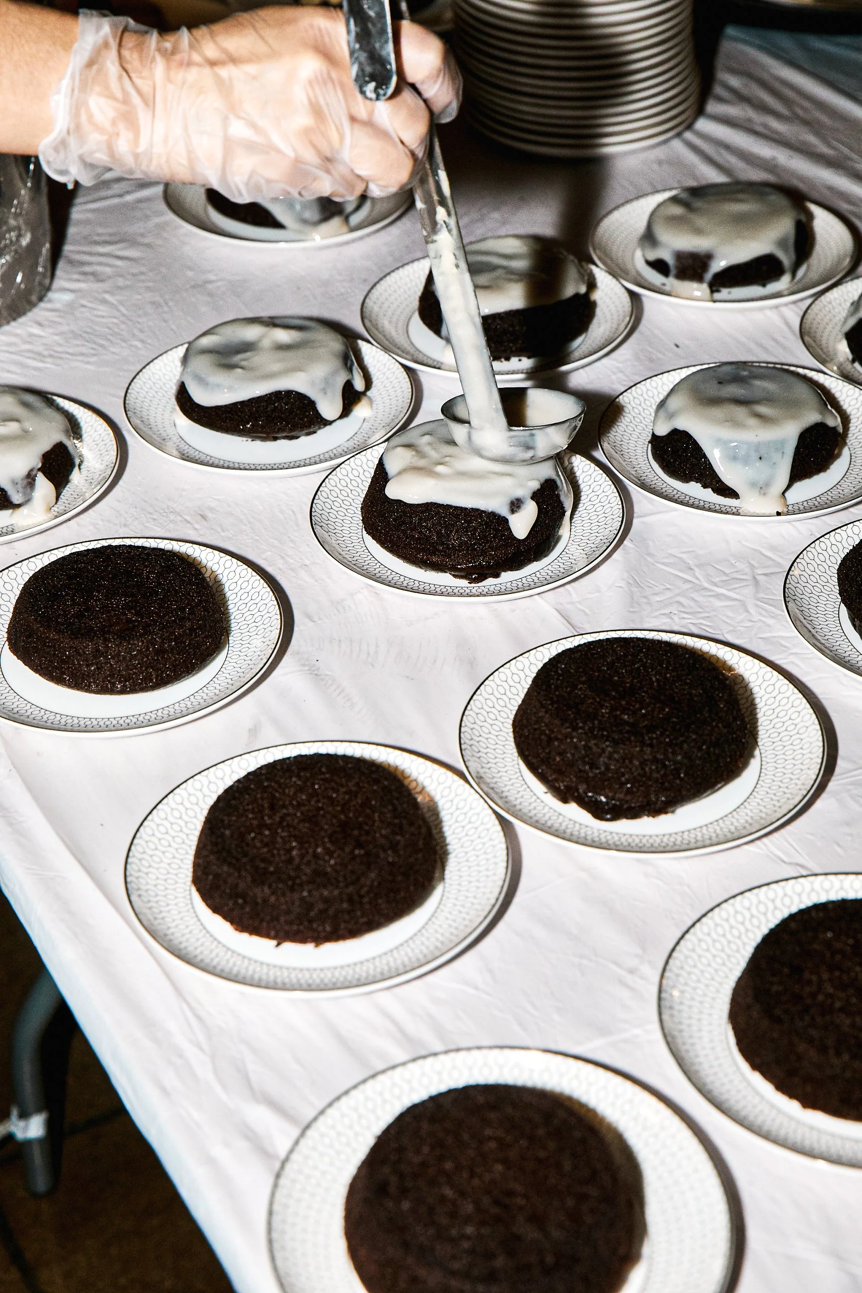 A person wearing gloves is pouring white icing or glaze over chocolate cakes on small plates, arranged on a white tablecloth.