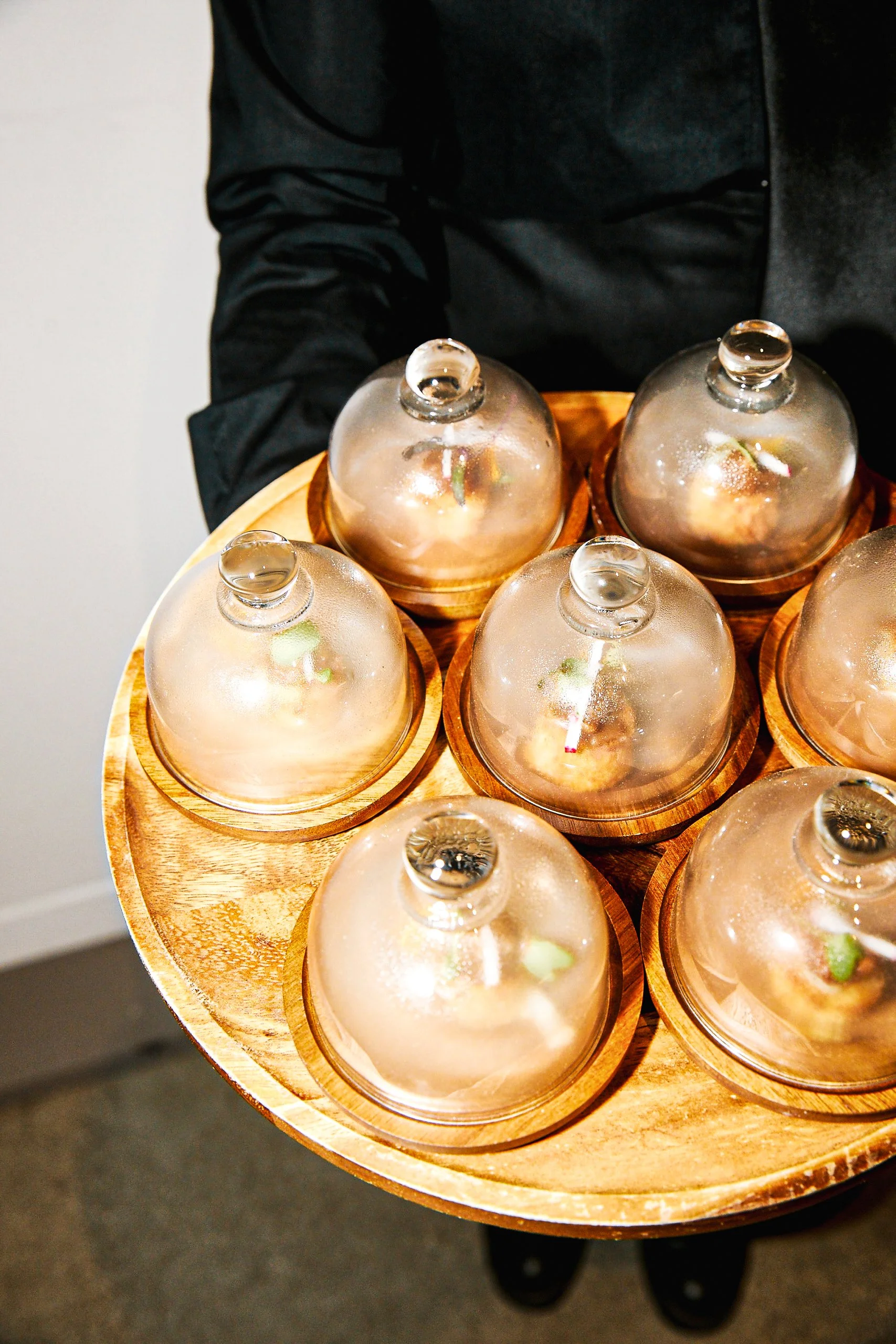 Person holding a round wooden tray with six glass cloches covering small food items, possibly desserts or appetizers.