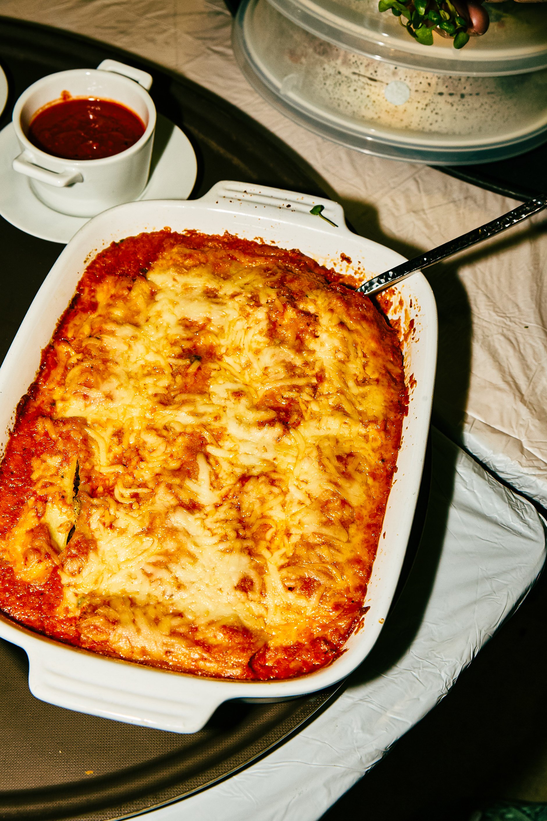 Baked lasagna with melted cheese in a white baking dish with a spoon, served with a cup of tomato sauce on the side.