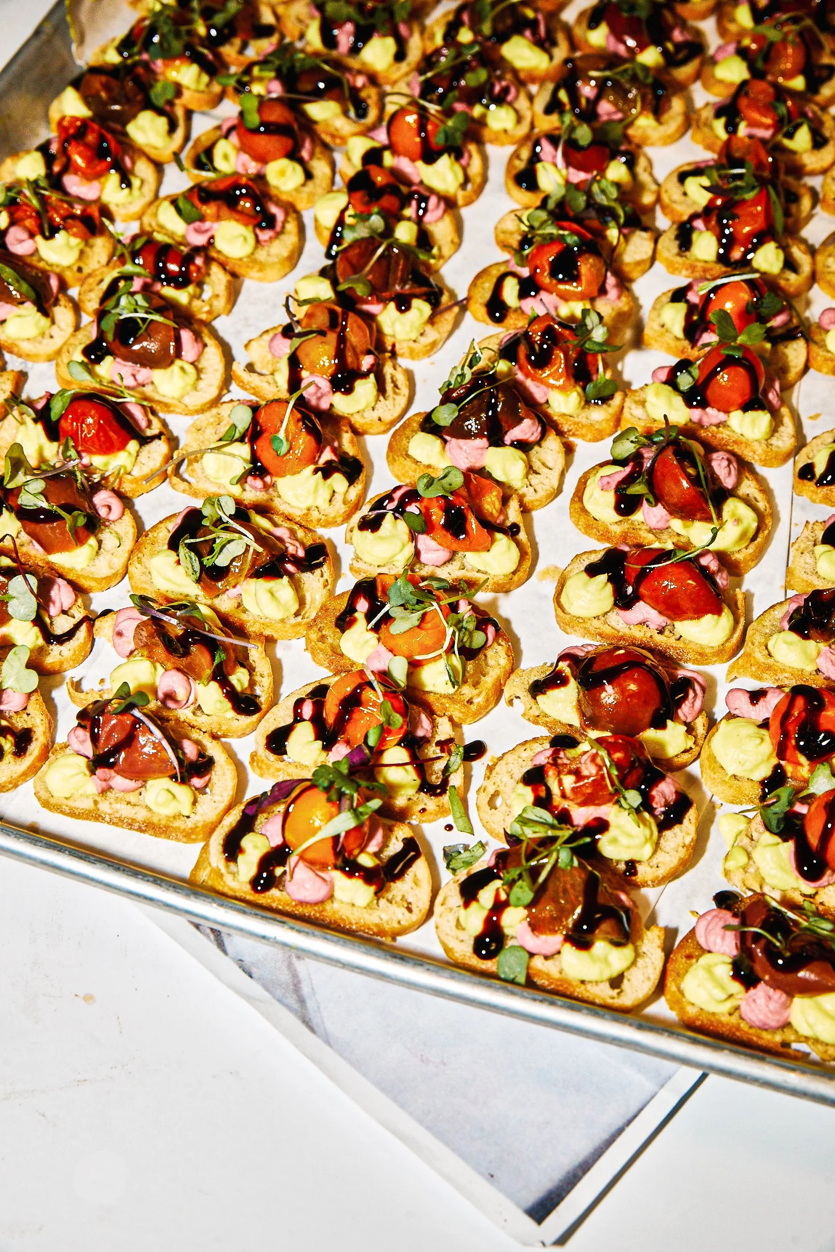 Assorted party crostini with cherry tomatoes, avocado, pink-colored toppings, microgreens, and drizzled balsamic glaze served on a tray.
