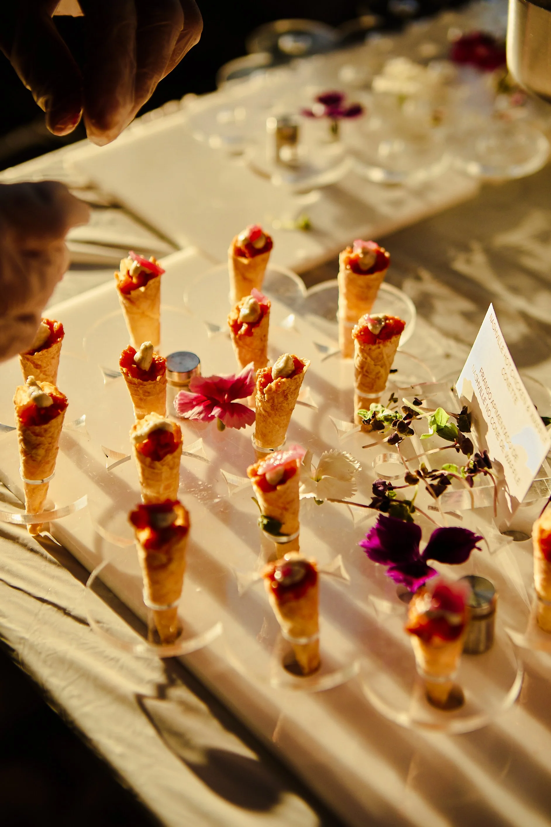 Multiple small ice cream cone desserts with toppings on a display table decorated with flowers at an outdoor event.