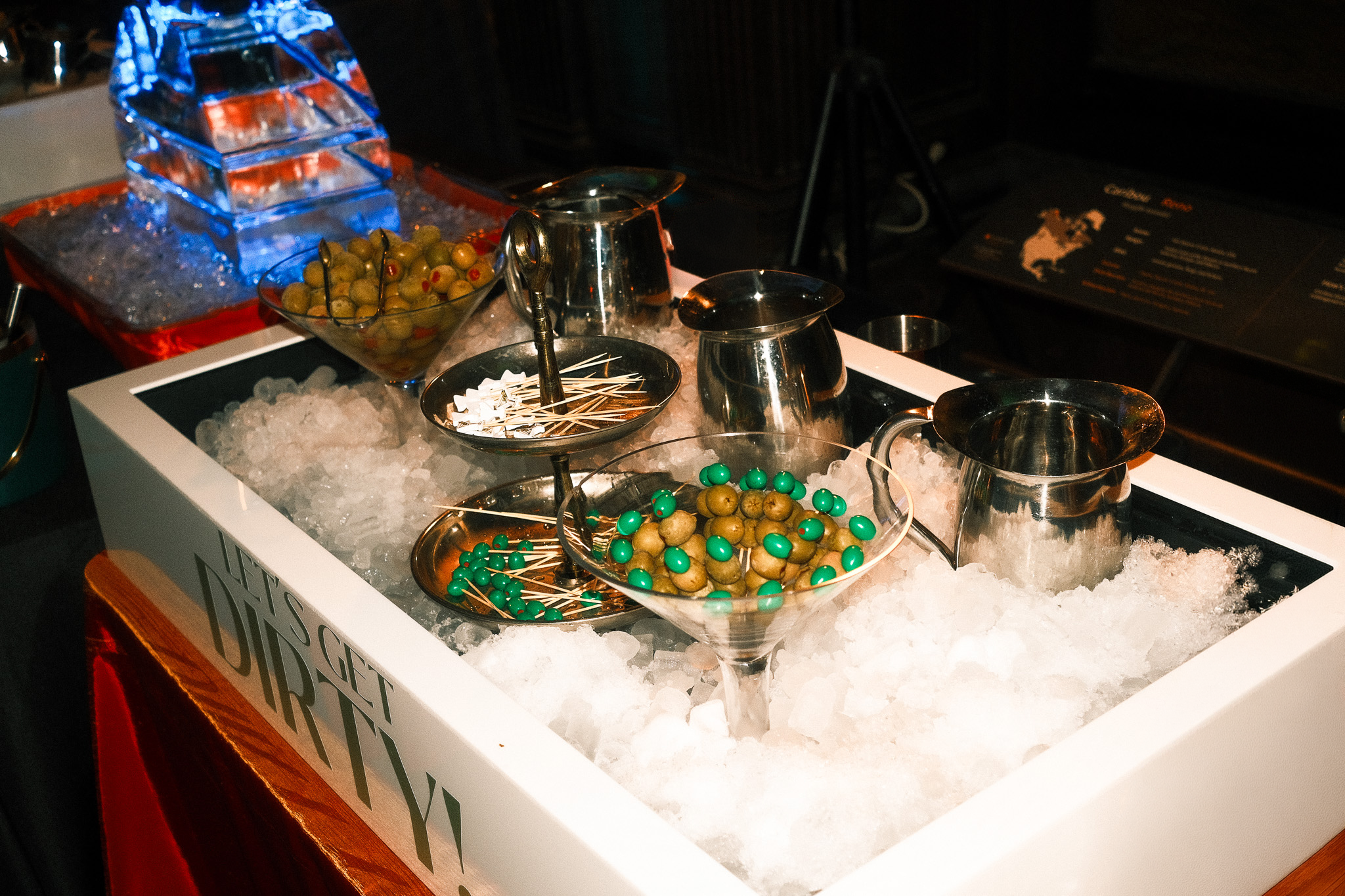 Ice bucket filled with ice, bowls of green olives with pimento stuffing and teal-colored candies on cocktail picks, and metal pitchers on a bed of crushed ice at a party or bar setup.