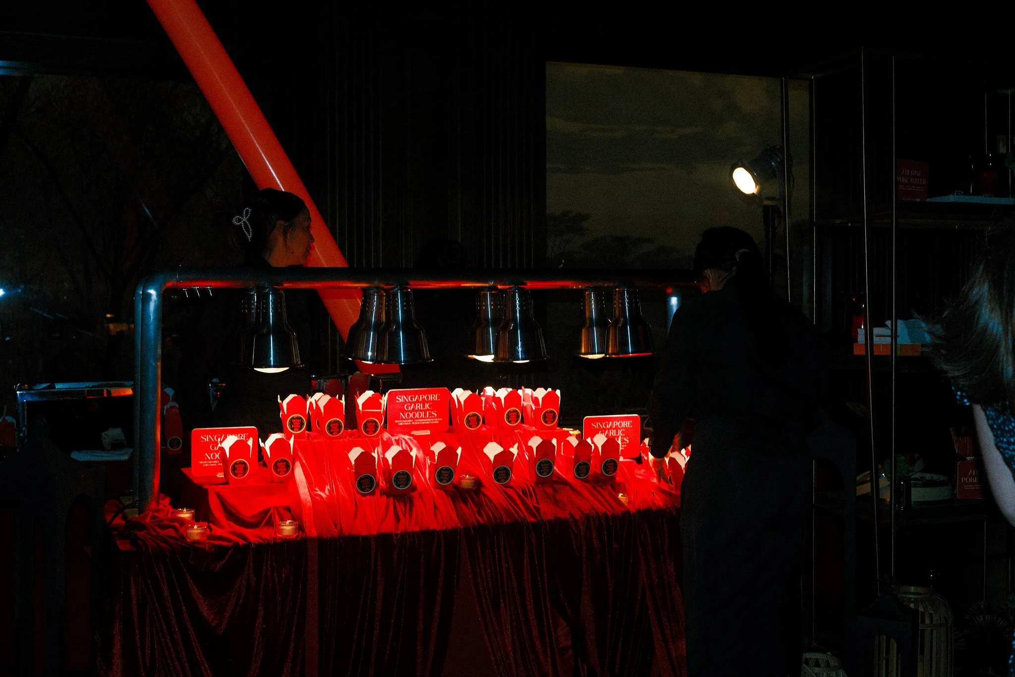 A dark indoor scene of a restaurant or food stall with a table decorated in red cloth, displaying takeout boxes labeled 'Singapore Garlic Noodles.' People are standing in front of the table, and there are lamps illuminating the boxes, with a window s