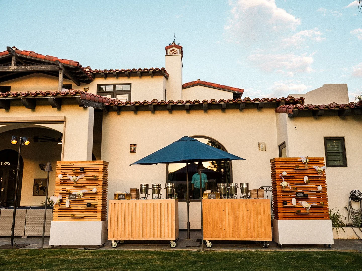 Outdoor event setup with a wooden bar, large blue umbrella, and decorative panels in front of a Mediterranean-style house with white walls and red tiled roof during daytime.