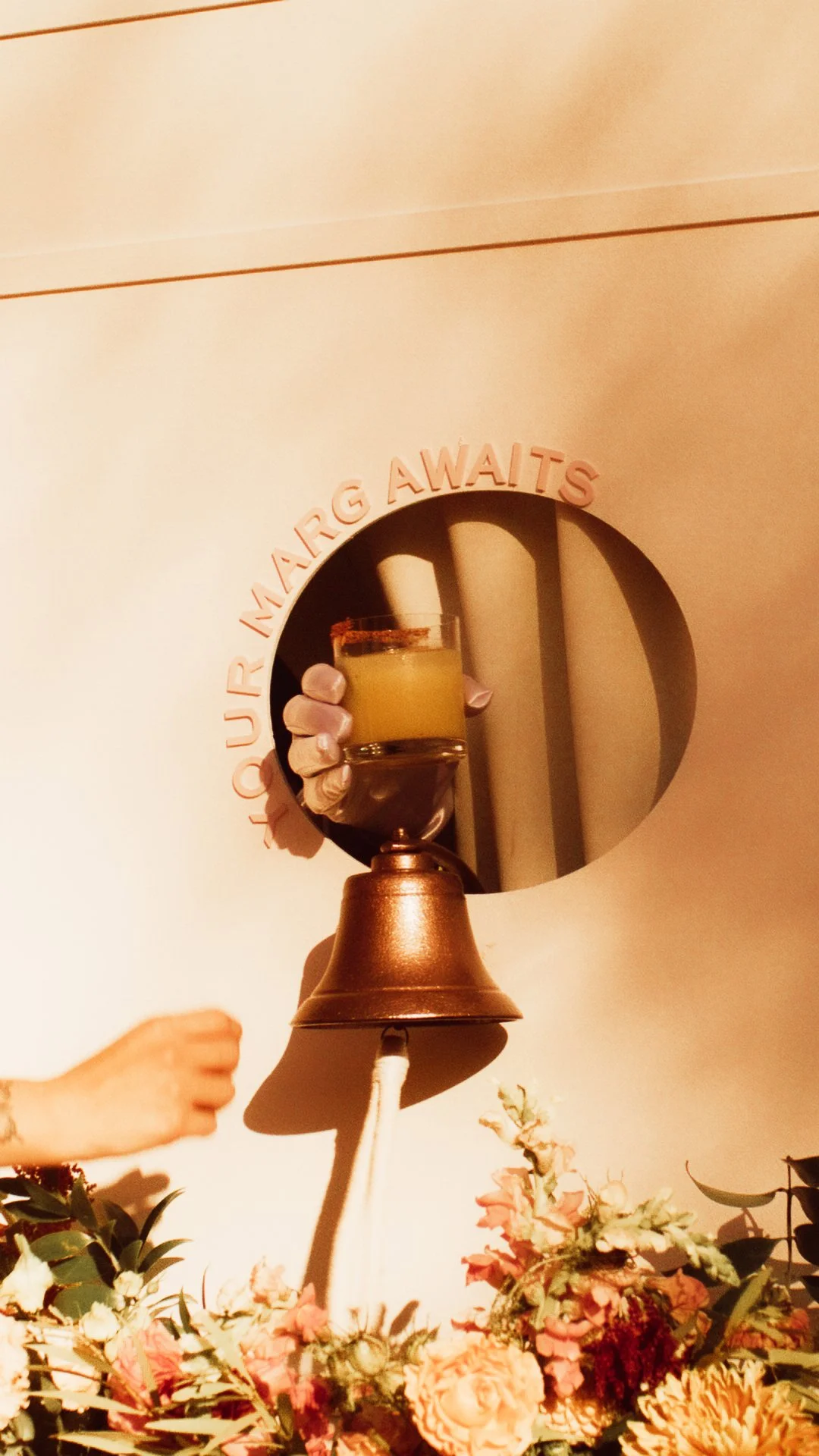 A hand holding a drink emerges from a hole labeled "Your MARG on ALERTS" in a white wall; flowers are in the foreground.