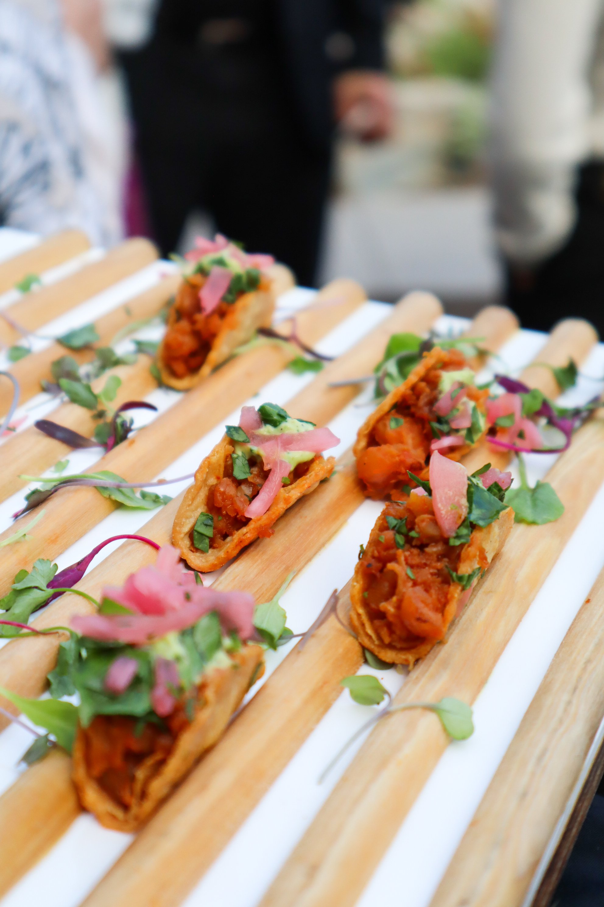 Close-up of mini tacos filled with meat, topped with chopped herbs and pink pickled onions, arranged on a wooden serving board decorated with small greens.