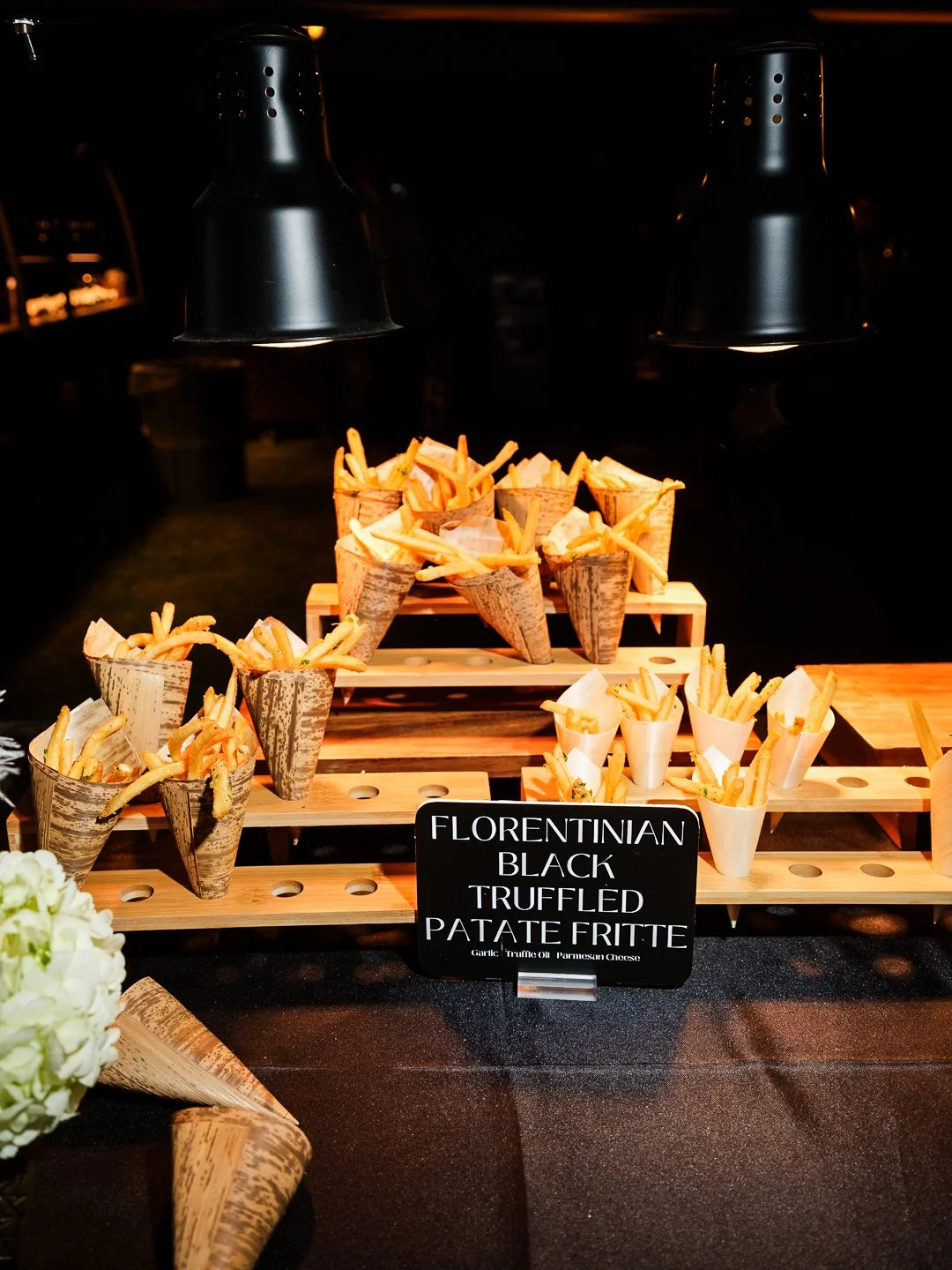 French fries served in cone-shaped paper wrappers on tiered wooden trays, illuminated by overhead lights at a buffet or catering event. A black sign indicates the fries are flavored with Florentine black truffle, accompanied by garlic, truffle oil, a