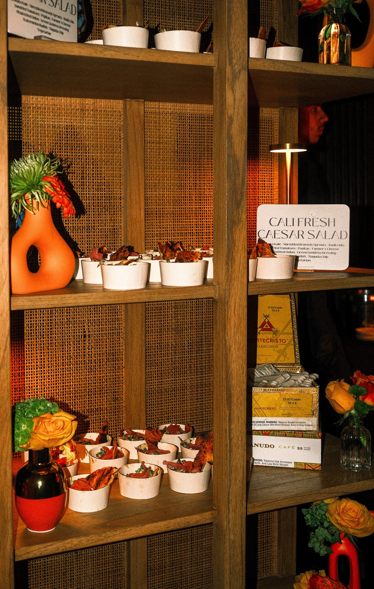 Display shelf with bowls of food, decorative vases, and flowers at a restaurant. A sign reads, 'CALI FRESH CAESAR SALAD' with a list of ingredients, and boxes of cigars are also visible.
