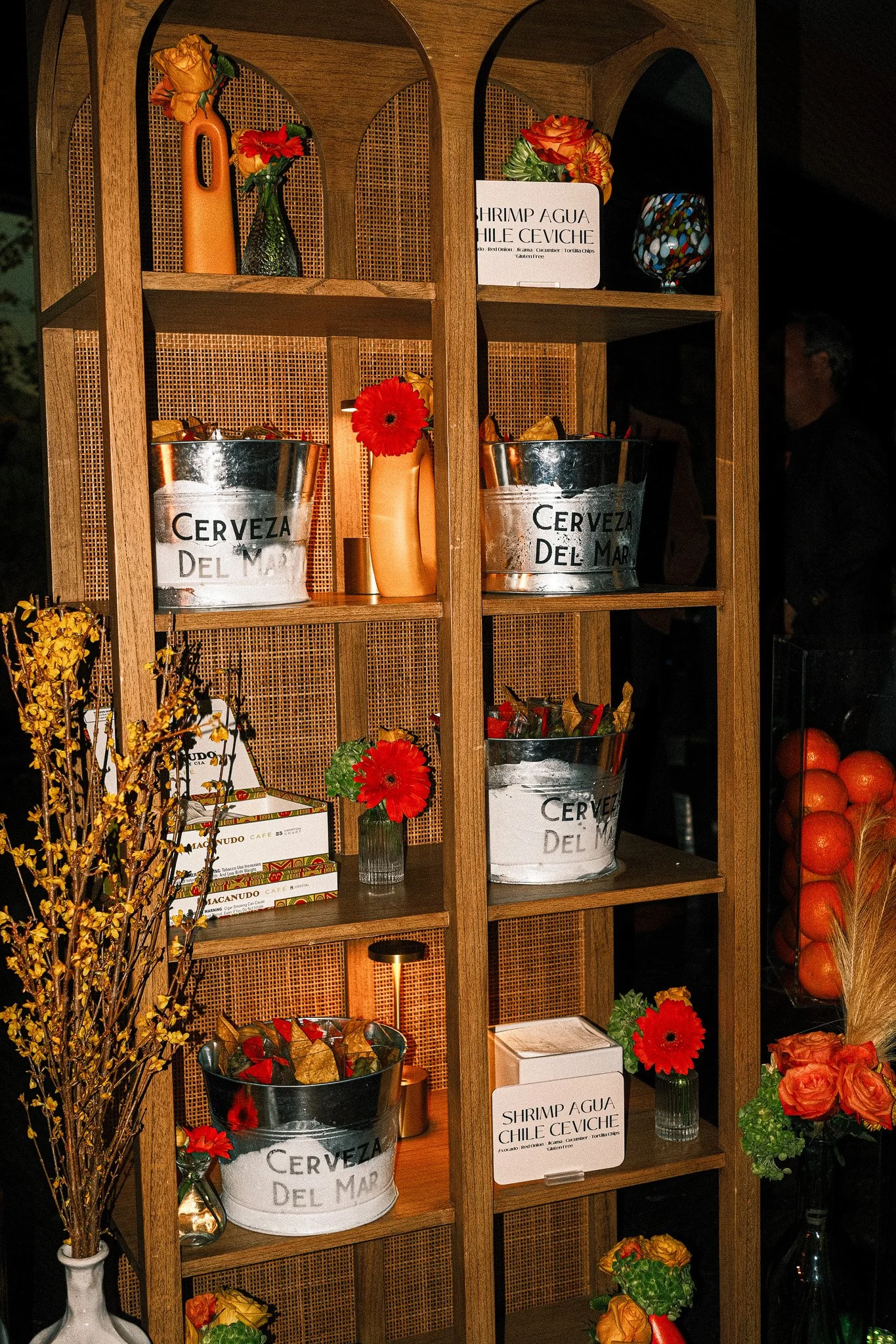Decorative wooden shelving unit with metal buckets labeled "Cerveza del Mar" filled with snacks and flowers, alongside small vases of flowers and signs for shrimp agua chile ceviche. Additional decorations include a tall vase of yellow flowers, orang
