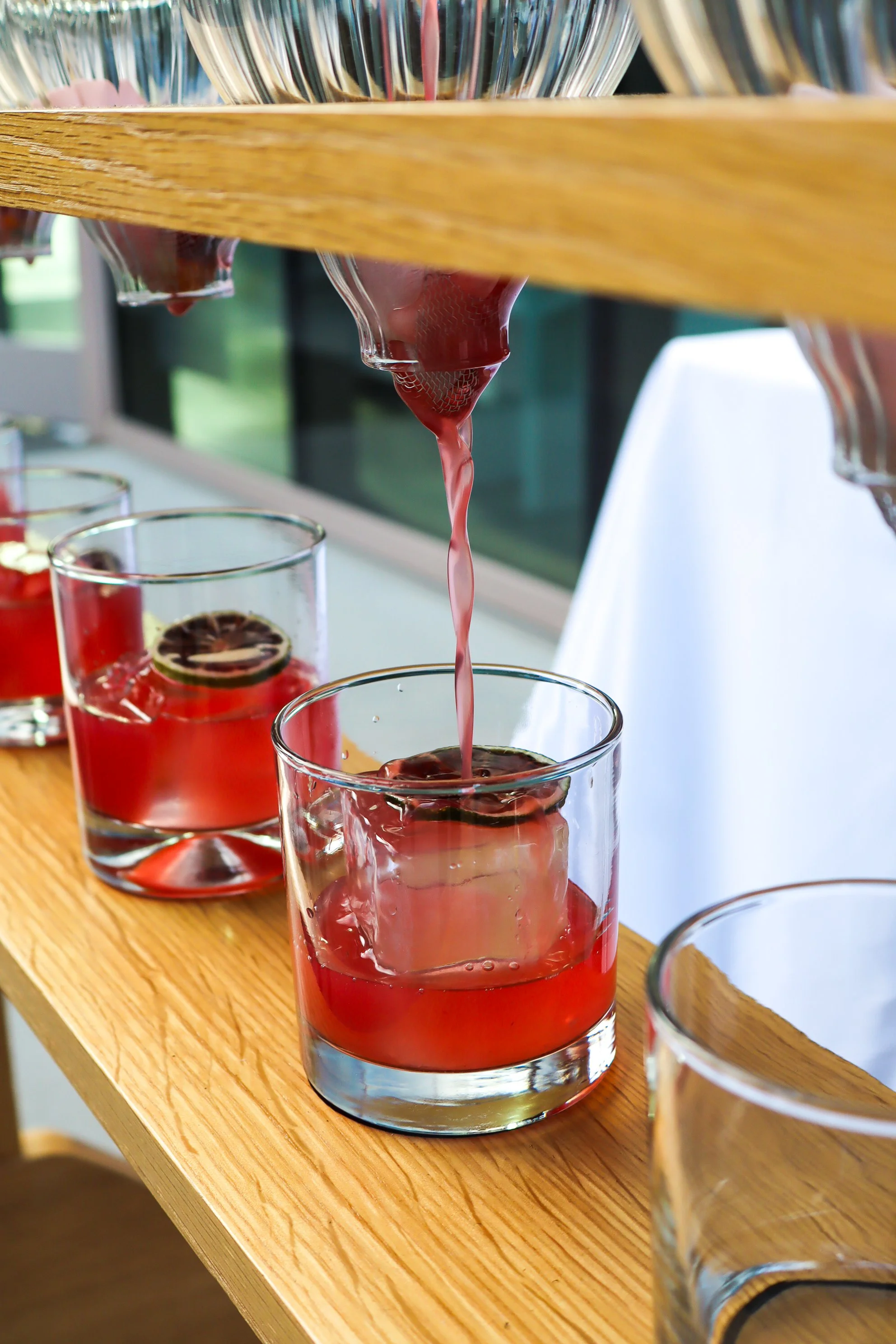 Glassware with red drink and lemon slices being poured from a glass container on a wooden table.