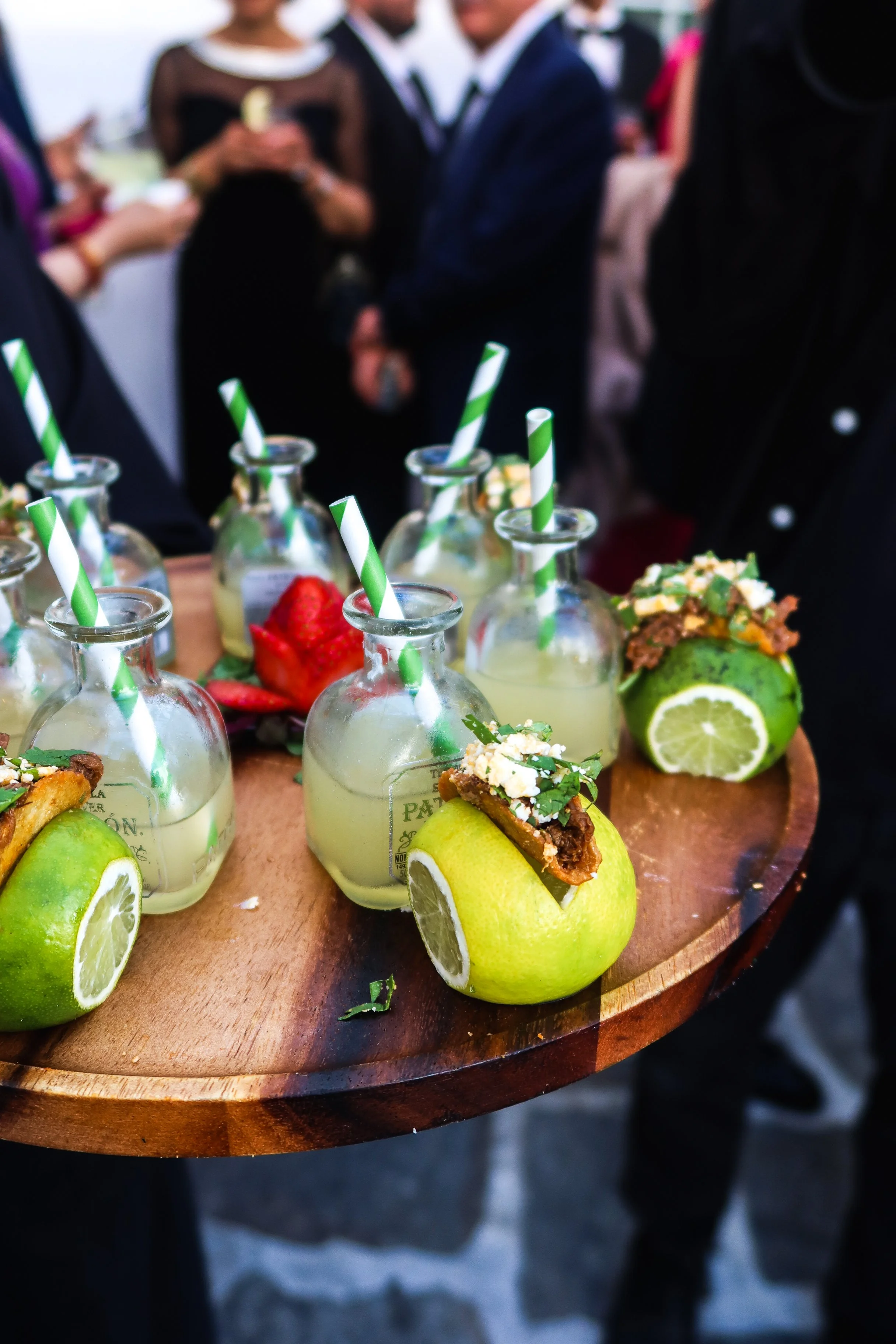 Wooden tray with green apples stuffed with a topping, glass bottles with lemon drinks and striped straws, and strawberries, held by a server at a crowded outdoor event.