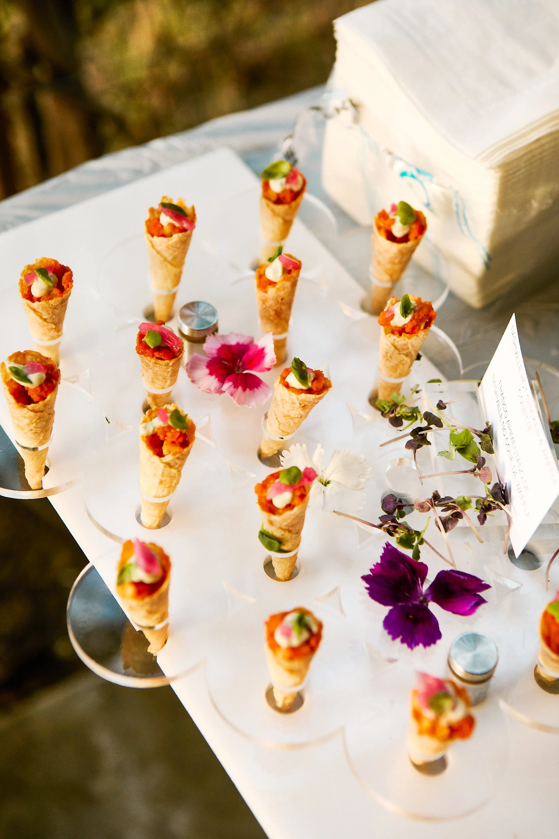 Miniature taco cones decorated with jalapeño slices and cream, displayed on a white stand with colorful flowers around.