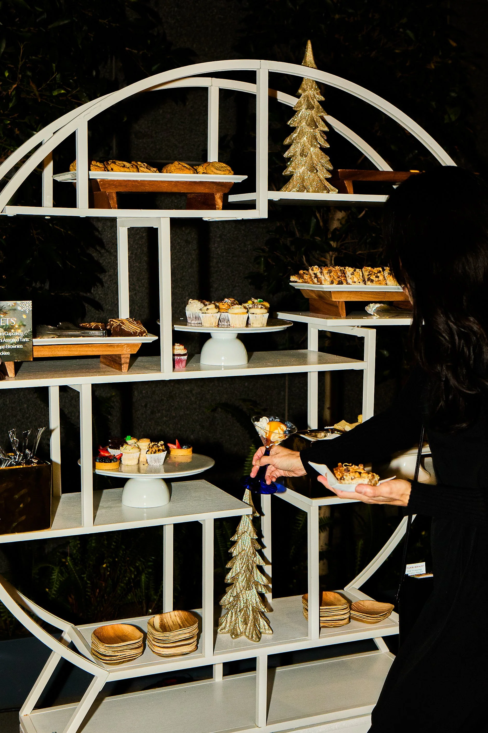 A woman selecting desserts from a multi-tiered white and wood display shelf at a holiday or festive event.