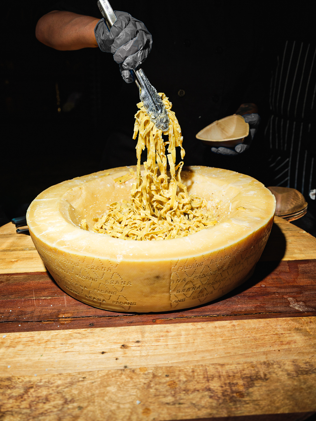 A person wearing black gloves scooping pasta from a large wheel of Parmesan cheese with tongs onto a wooden table.