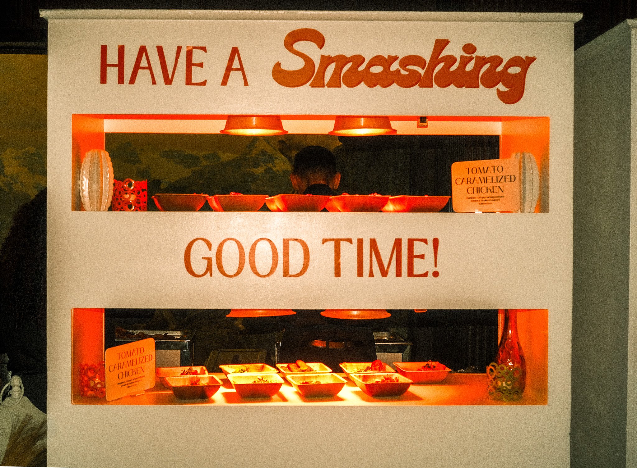 A food stand with the sign 'Have a Smashing Good Time!' featuring bowls of food and a small sign labeled 'Tomato Caramelized Chicken'.
