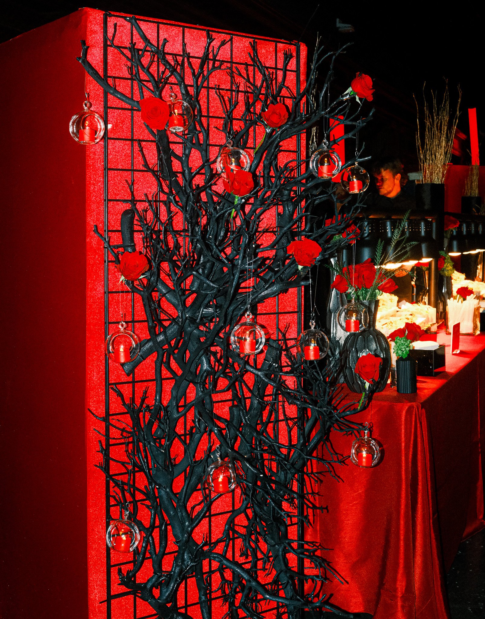 Decorative black tree with red roses and hanging glass orbs, placed against a red textured background, part of a red-draped display table with flowers and candles, in a dimly lit setting.