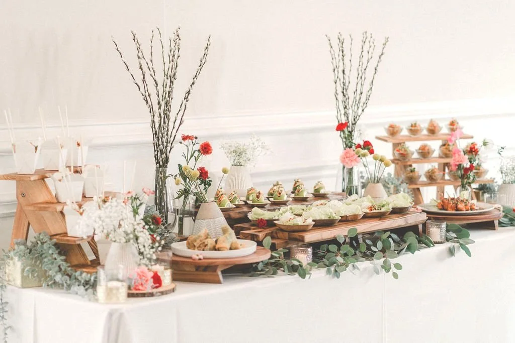 Drop-off catering, Long beach, dessert table decorated with flowers, greenery, and wooden serving trays, featuring cupcakes, cookies, and other treats.