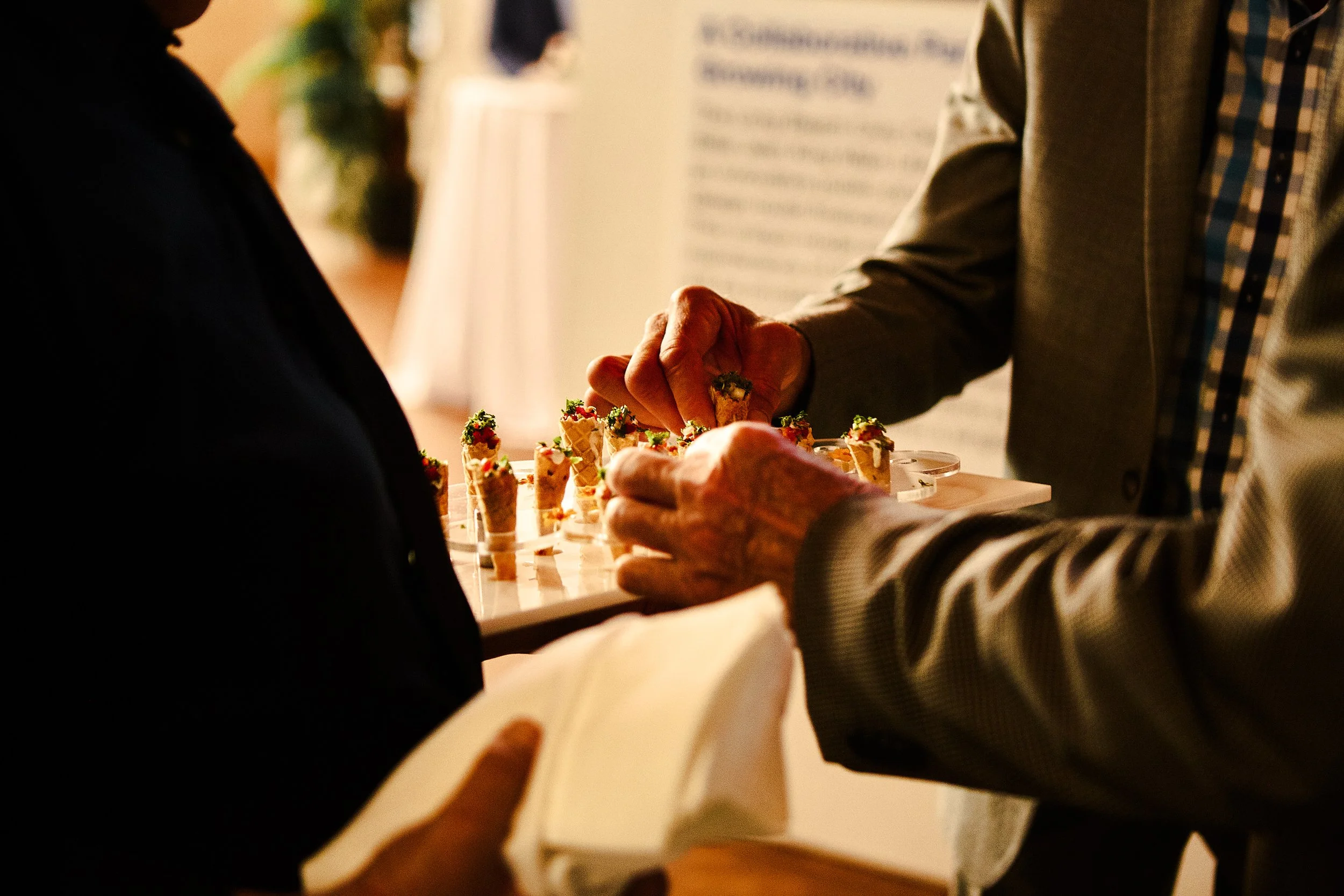 Two people serve appetizers or desserts at a social event, with one person holding a plate and the other selecting small food items from a tray.