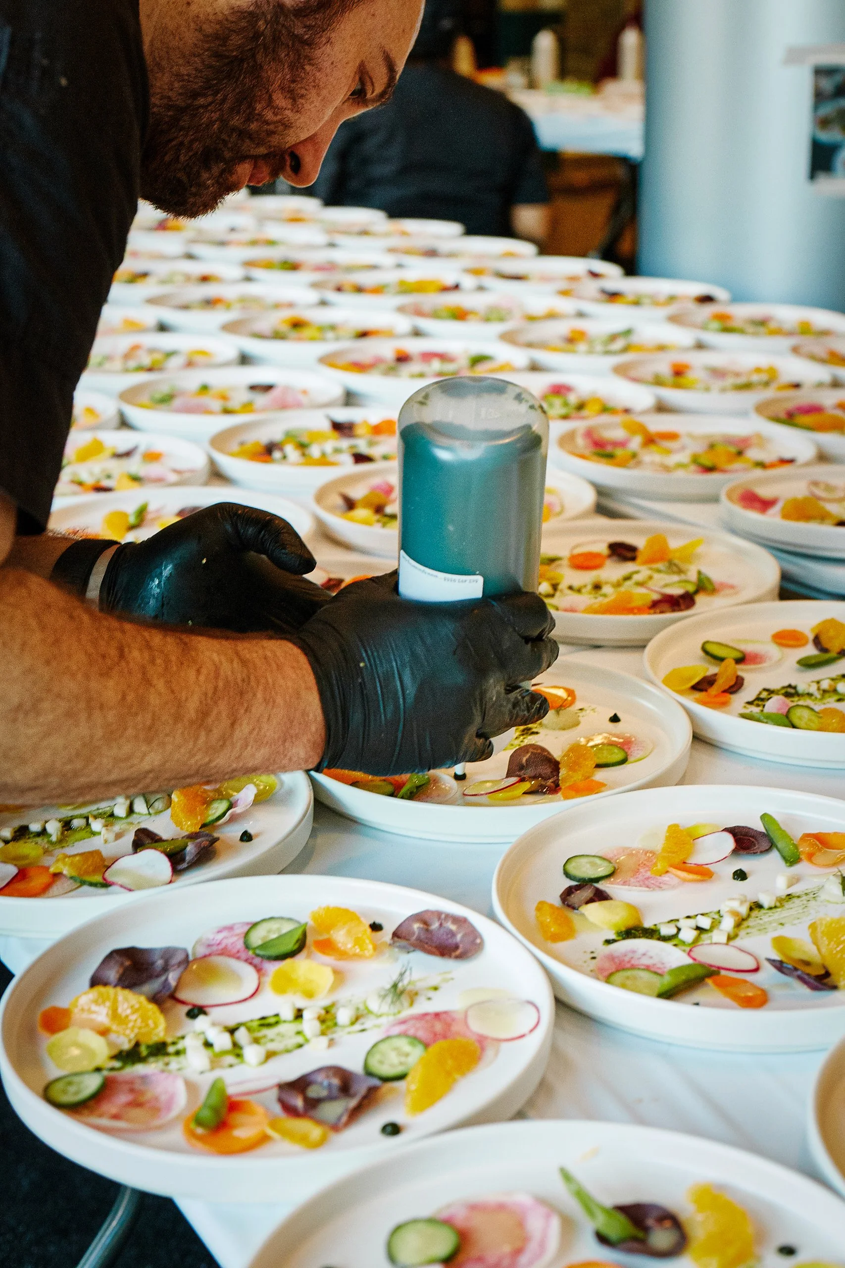 A chef with black gloves is decorating plates with food, surrounded by numerous plates of colorful food with sliced vegetables and garnishes.