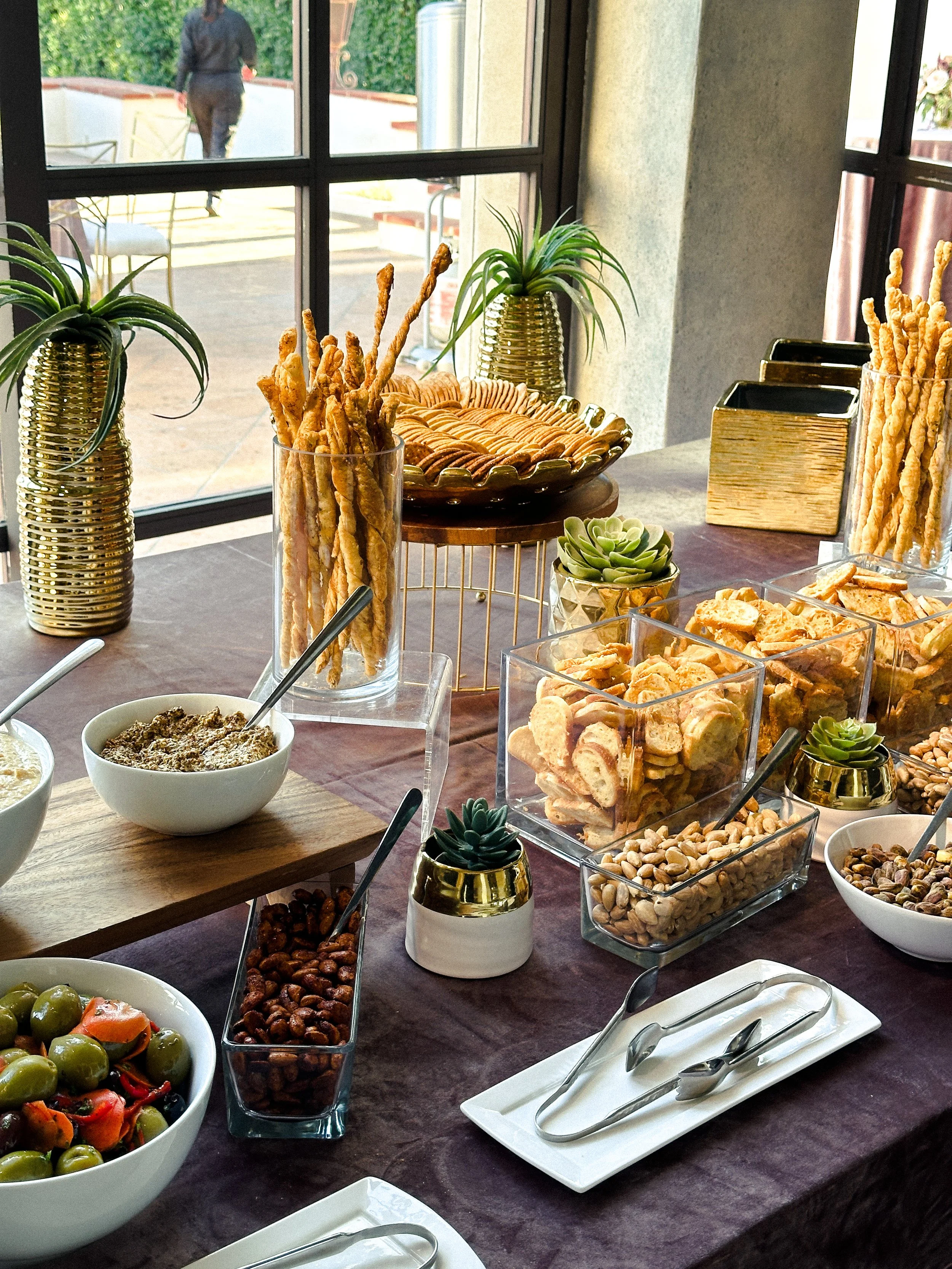 A snack table with bowls of peanuts, chips, and olives, and decorative plants near a window.